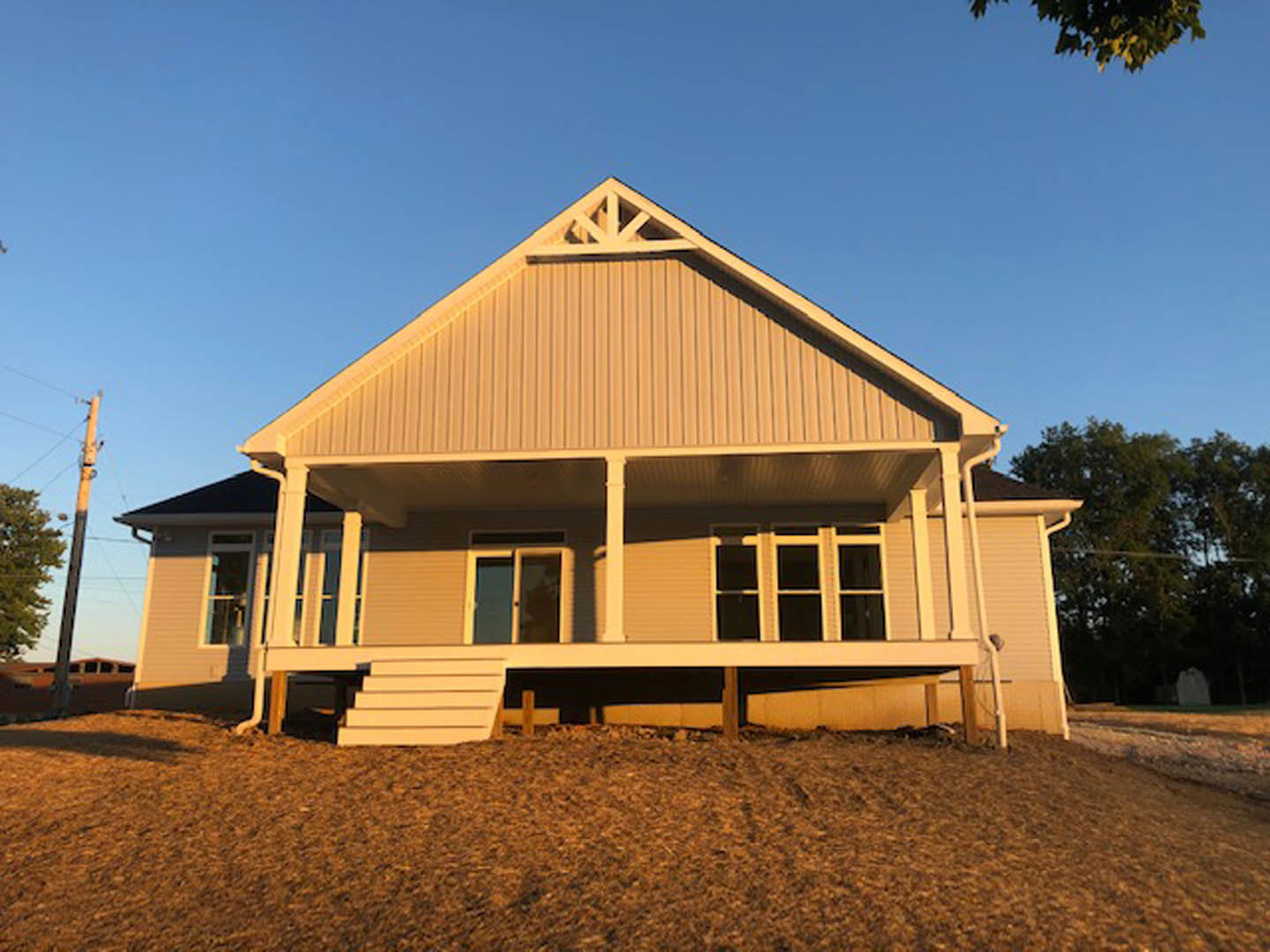 Front porch with white railings, light-colored siding, white-framed windows, brown dirt yard, wooden fence, and trees in the background