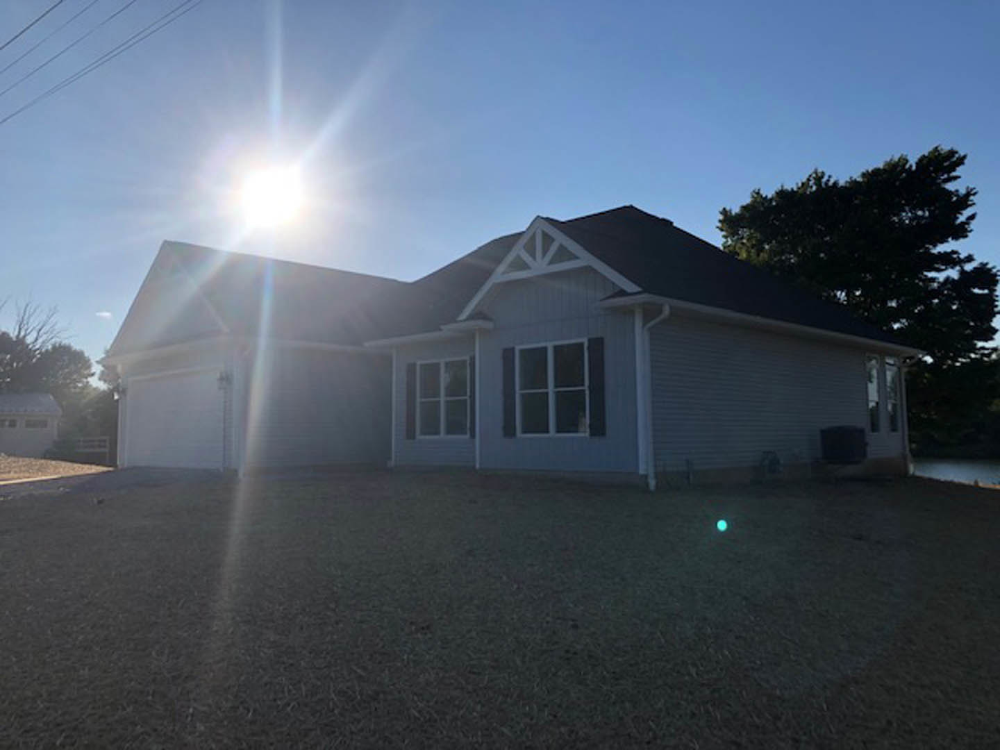 Two-story house with attached garage, white window frames, light-colored siding, green lawn, and mature tree in the background under a clear blue sky