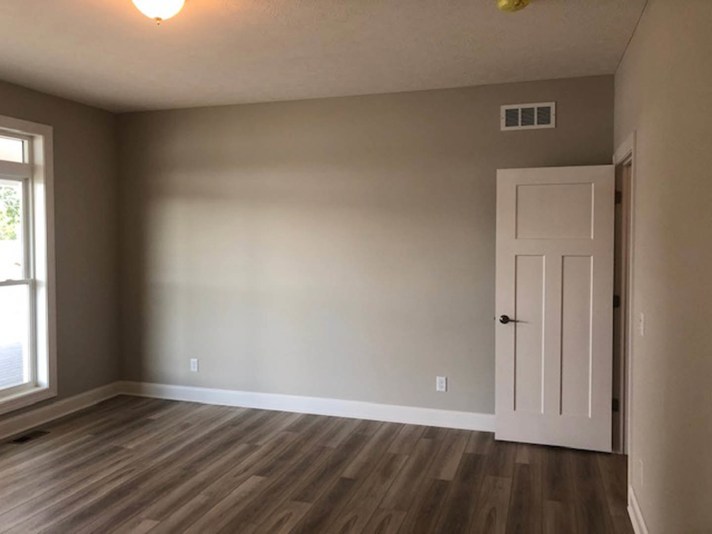 Hardwood floor room with white door featuring black handle, white walls, ceiling light fixture, window with vent, and plaster ceiling.