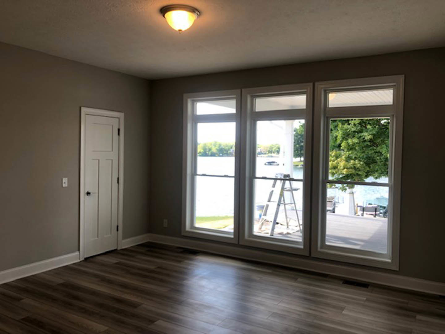 Wood floor with sunlight, white walls, black-handled white door, ceiling light fixture, window with ladder leaning outside