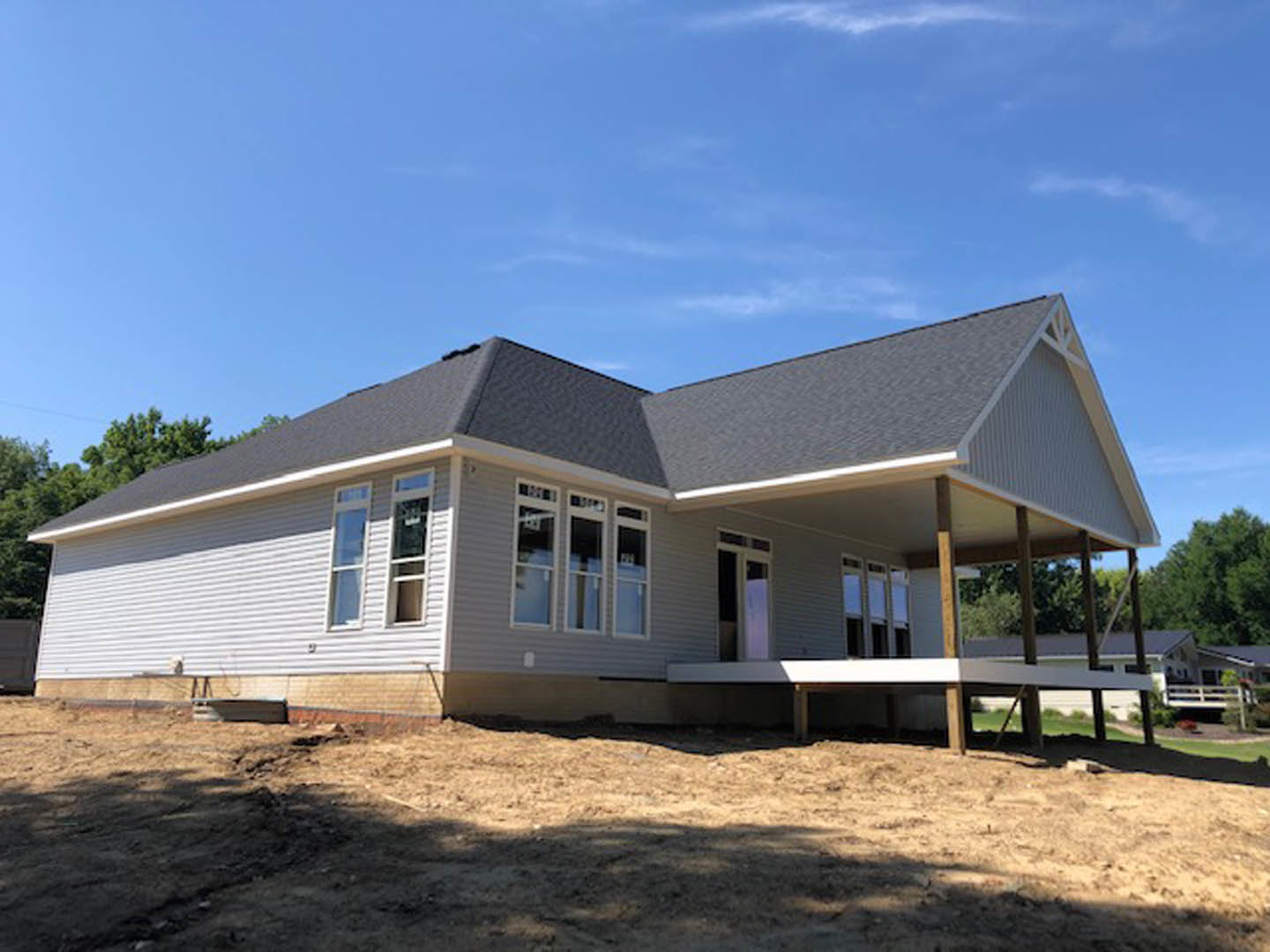 Wood-framed house under construction with covered porch, exposed beams, unfinished siding, dirt yard, and large windows beneath blue sky