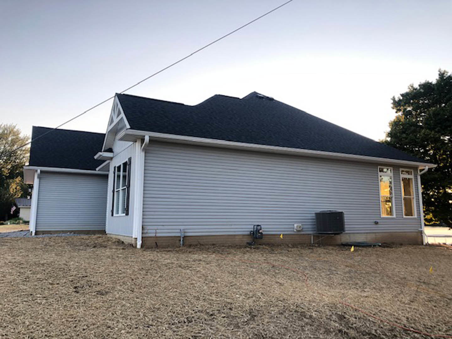 Two-story home with gray siding and white-trimmed windows, dark shingle roof, spacious backyard with patchy brown grass, metal fence, and trees under a clear sky