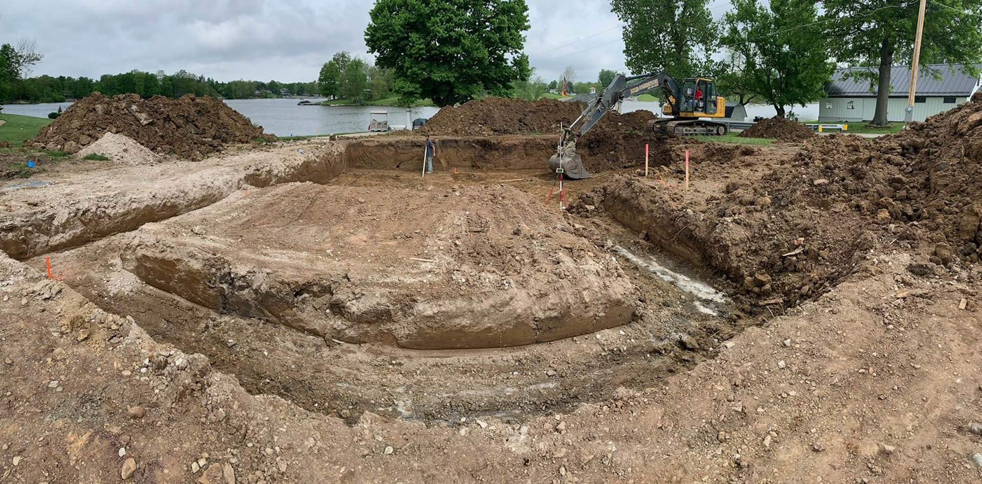 Bulldozer parked beside large mound of soil on residential construction site, surrounded by muddy ground and leafy trees under cloudy sky