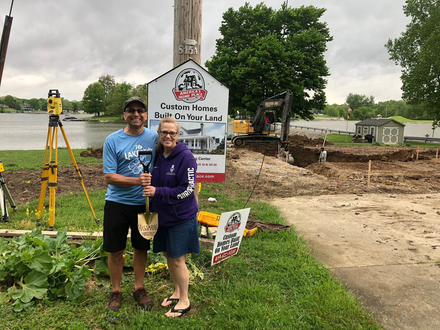 Couple standing on grassy lawn beside a custom home construction sign, man holding shovel, woman smiling, trees and cloudy sky in background