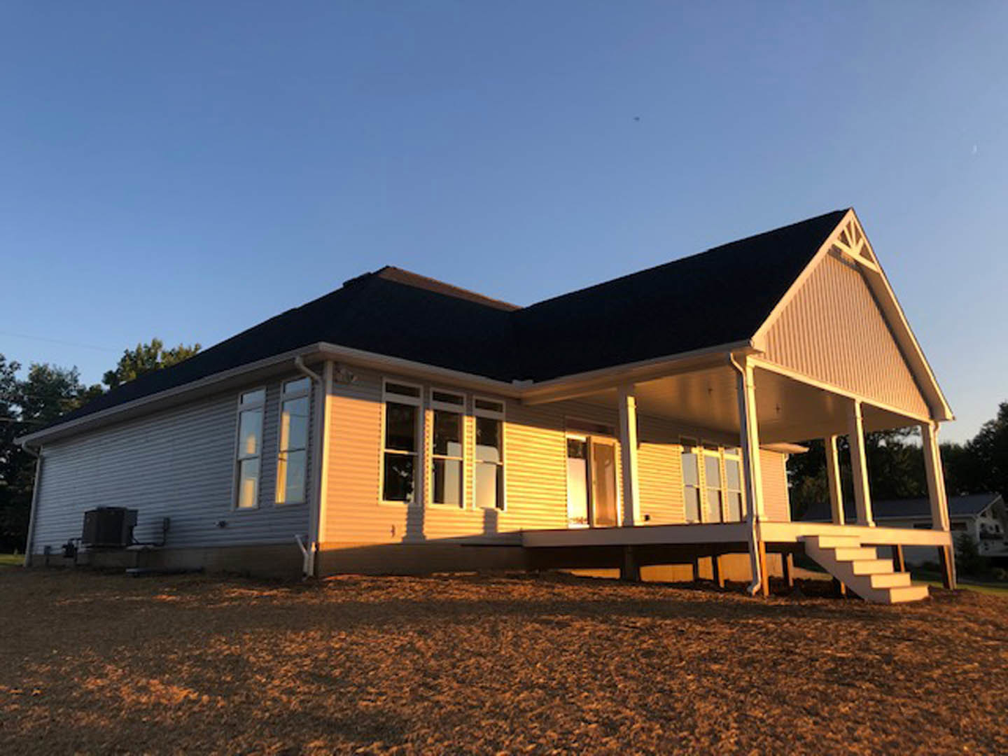 White siding house with covered front porch, white columns, large windows, manicured lawn, leafy tree, and clear blue sky