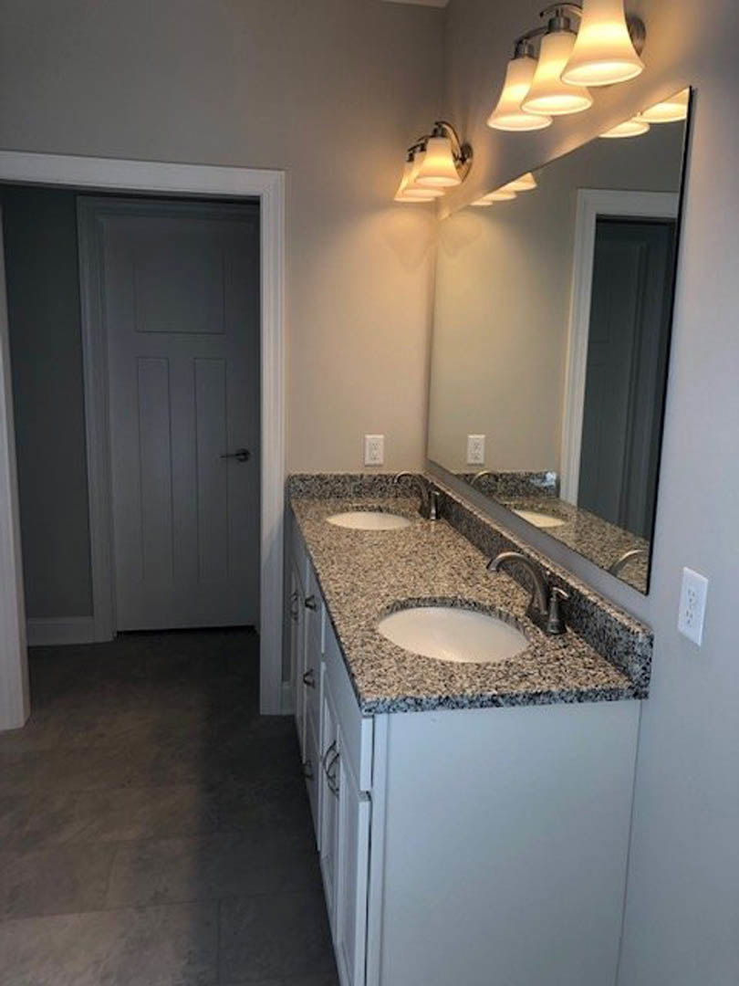 Bathroom with double vanity featuring undermount sinks, wide framed mirror, row of wall-mounted lights above, grey tile flooring, white cabinetry, and a door with black hardware.