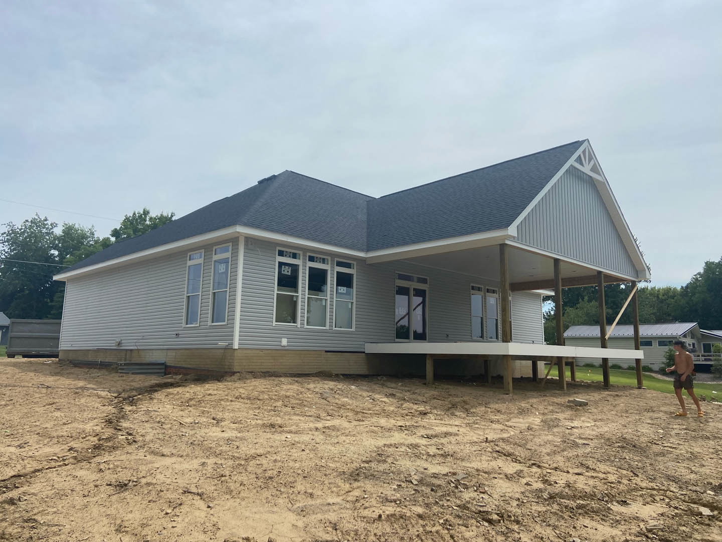 Two-story house under construction with exposed framing, white window frames, covered porch, dirt yard, and surrounding trees