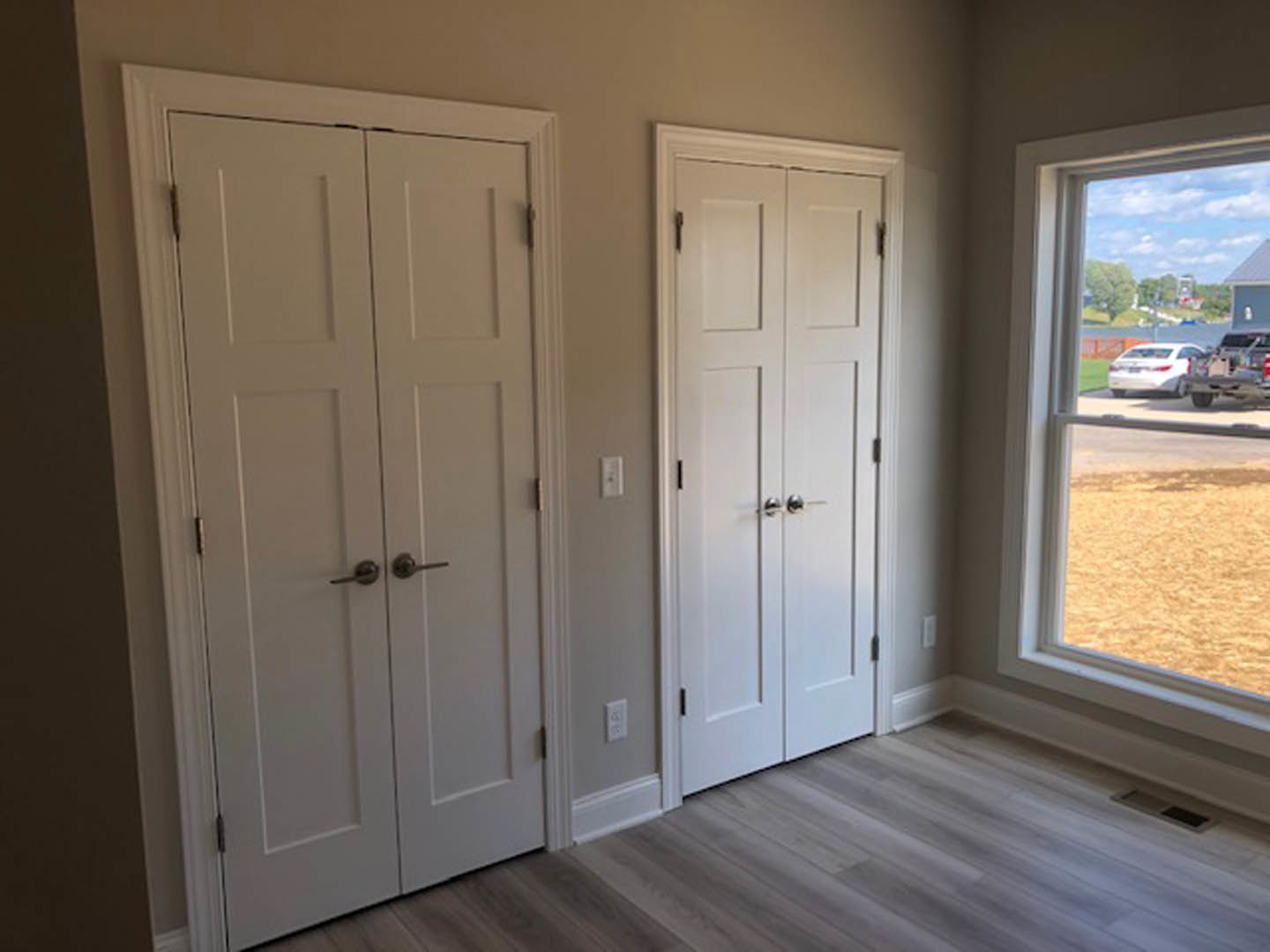 White-painted room featuring two doors with silver handles, a window overlooking a parking lot with cars, wood flooring, and neutral walls.