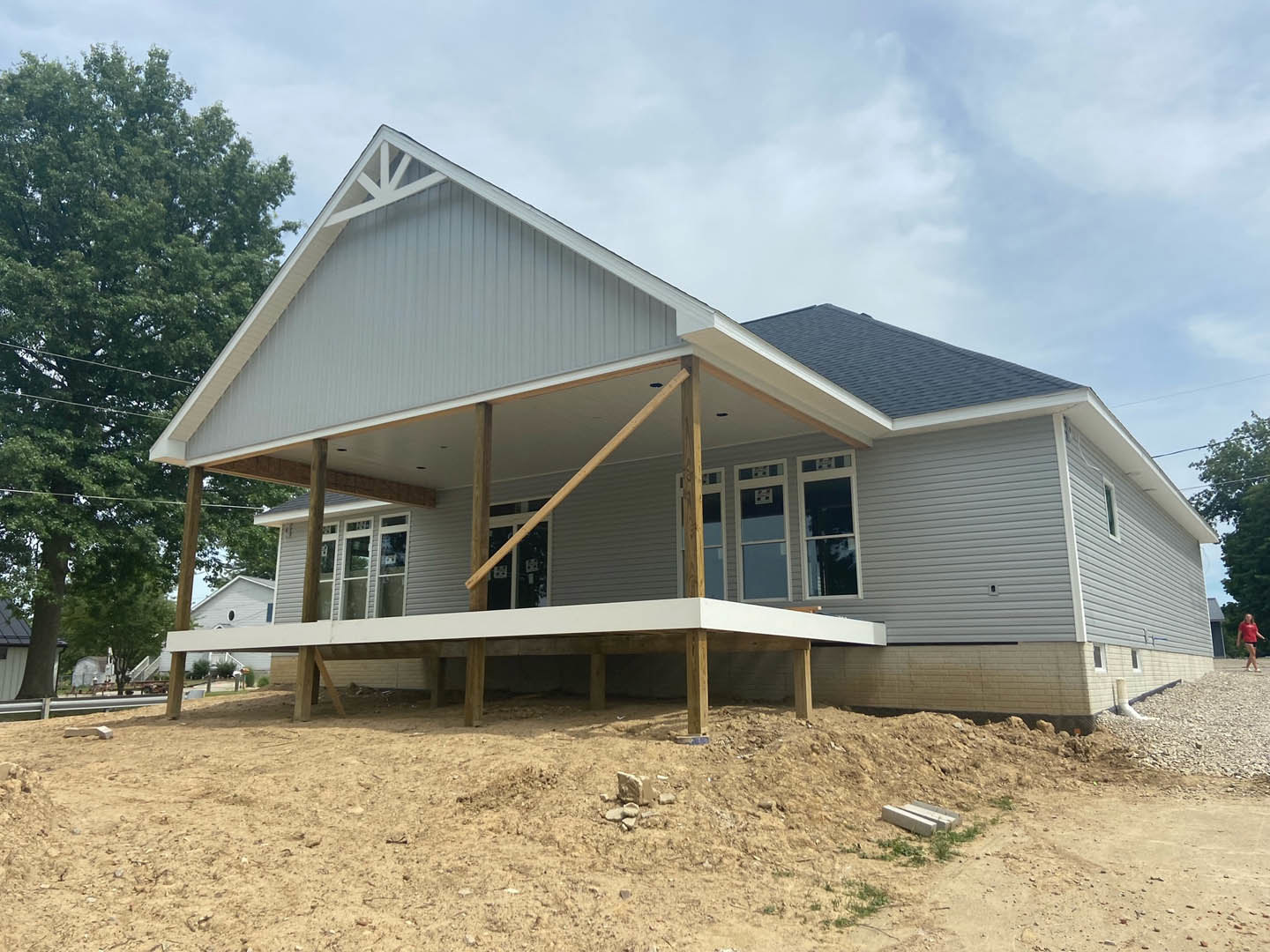 Partially built house with white-framed window, exposed white siding, unfinished porch, dirt yard, and pile of rocks; tree and cloudy sky in background