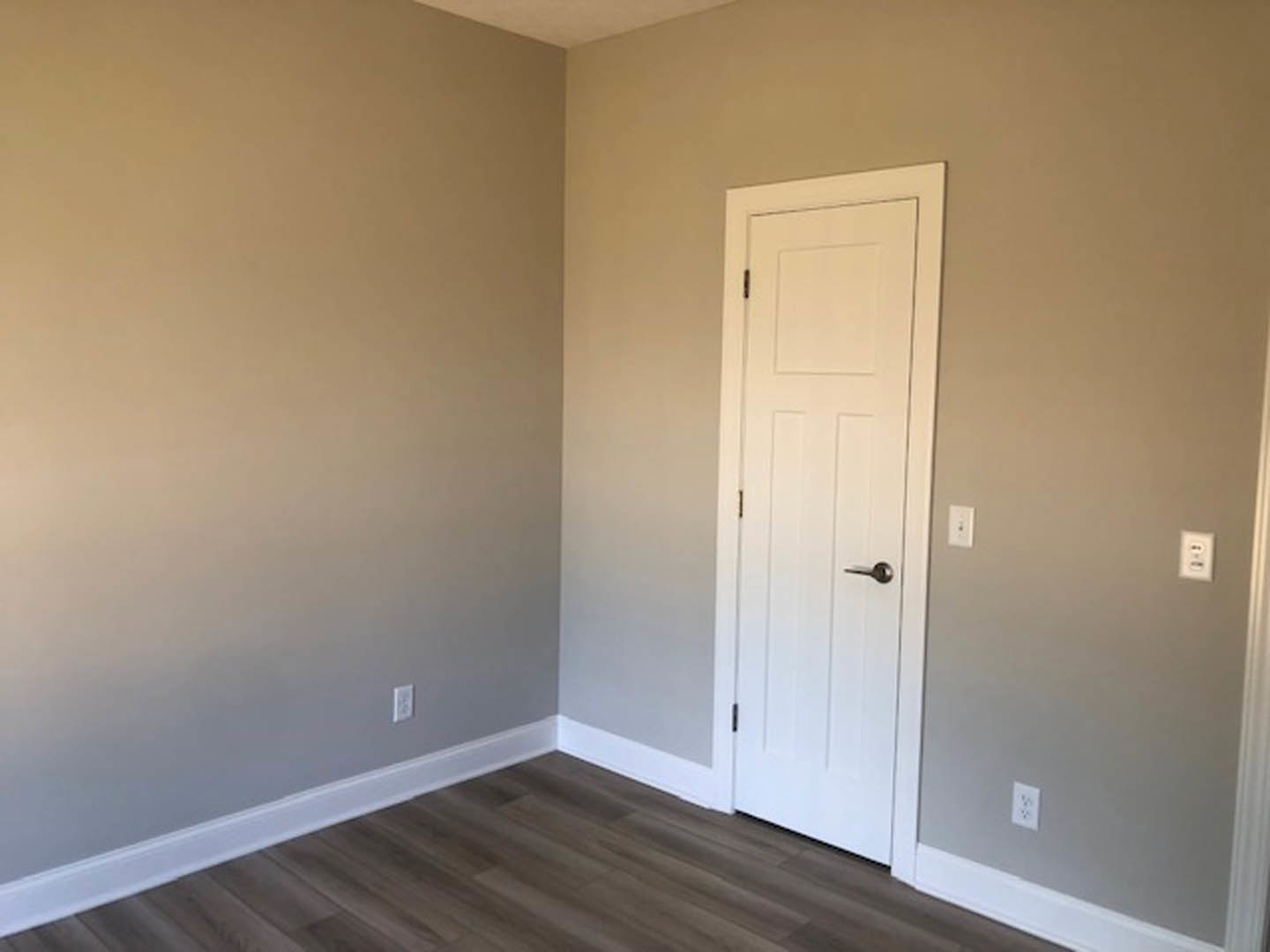 White paneled door with silver handle, wood floor with white baseboard, wall outlet on light-colored wall