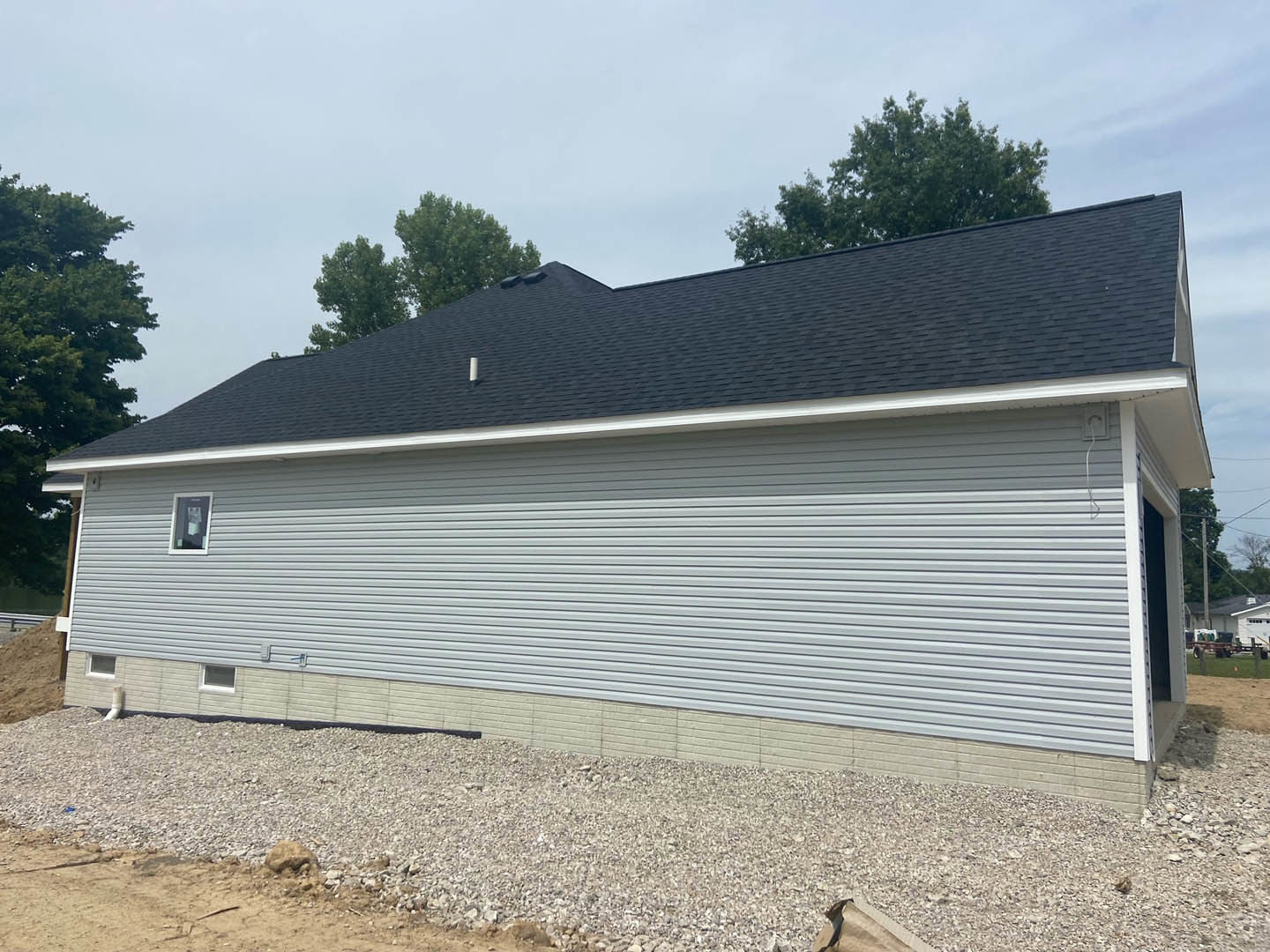 Modern house with black roof, gravel front yard, mature trees, light-colored exterior walls, large window displaying a sign