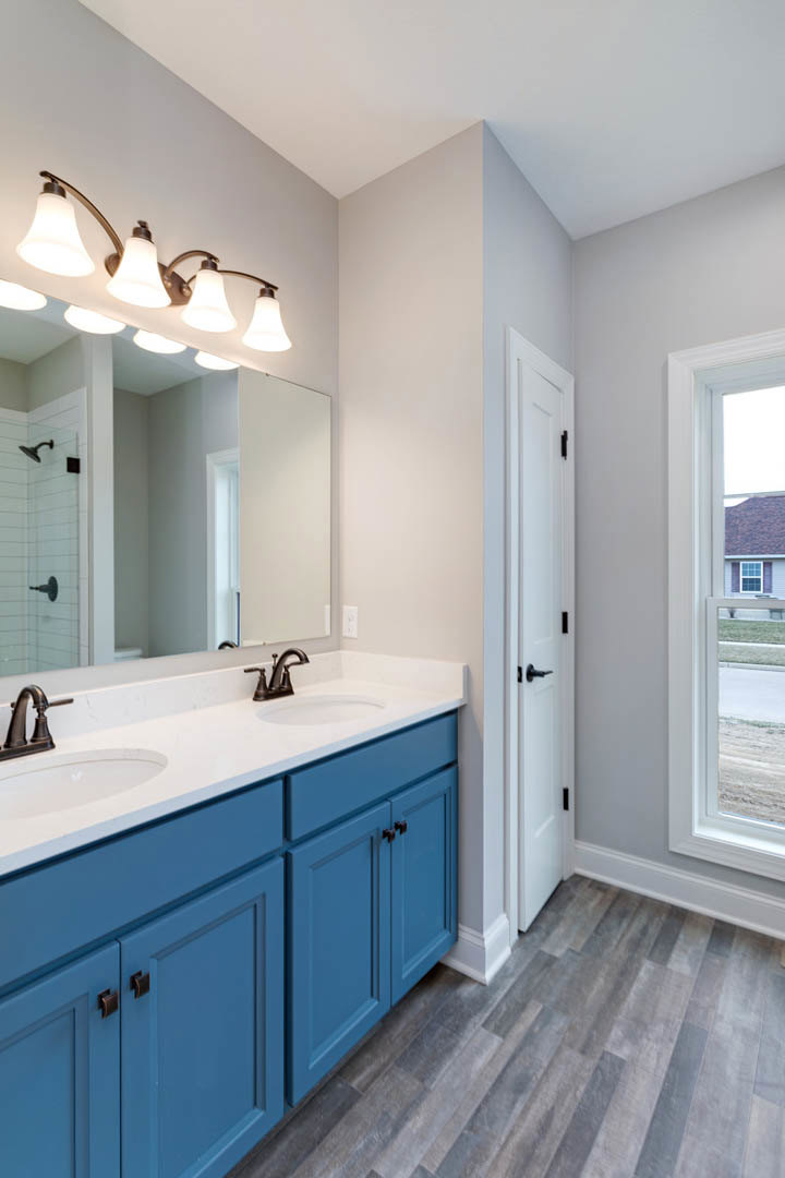 Bathroom with blue shaker cabinets, quartz countertop, rectangular mirror above sink, chrome faucet, row of globe light fixtures, white door with black handle, window showing