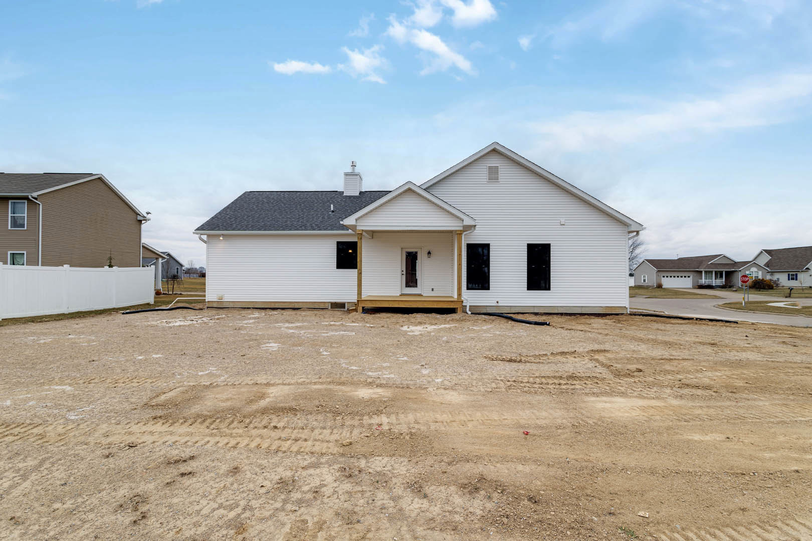 White house with front porch, black door framed in white trim, dirt area with visible tire tracks in foreground, cloudy sky overhead