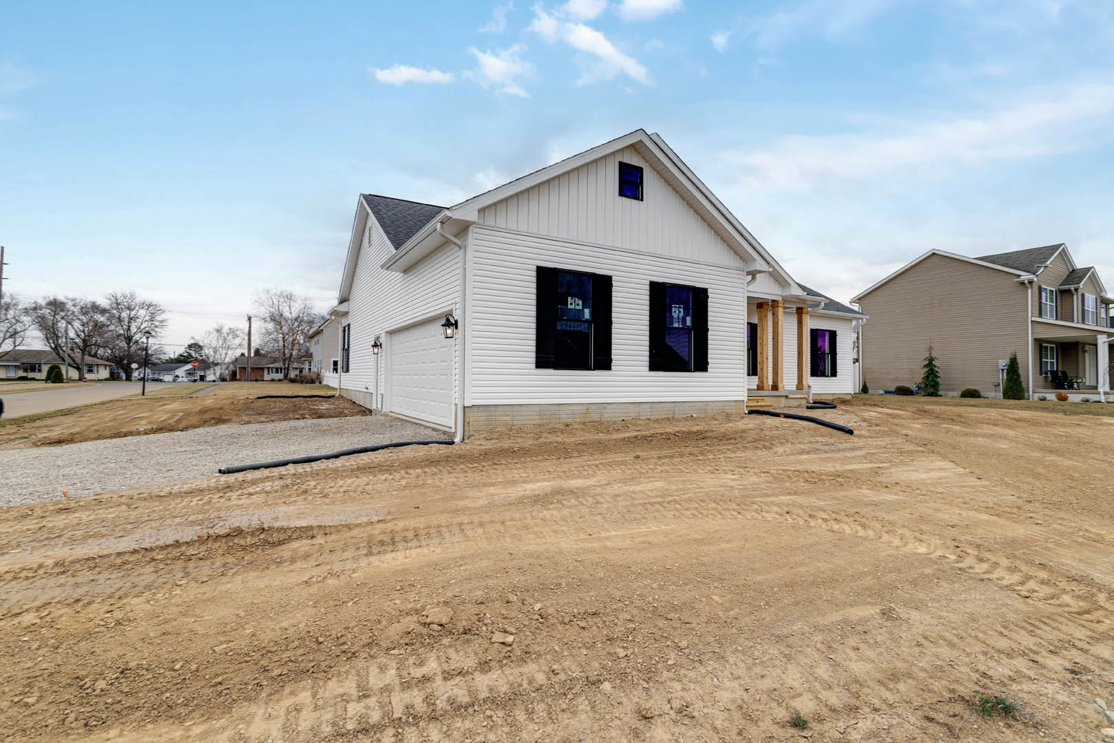 White house with black-framed windows, dirt road with tire tracks leading to the entrance, sparse trees around the property, partly cloudy sky overhead