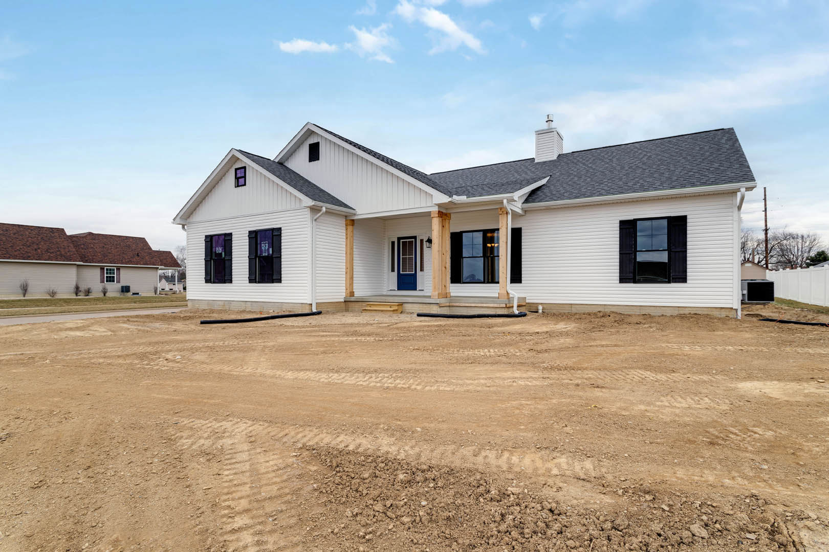 White house with blue door and red roof, dirt yard with tire tracks, multiple windows, cloudy sky above