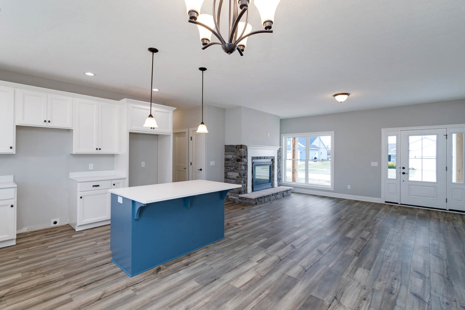 Open-concept kitchen and living room featuring a blue and white island countertop, white cabinetry, stone fireplace, hardwood floors, glass-paneled door, large windows with outdoor