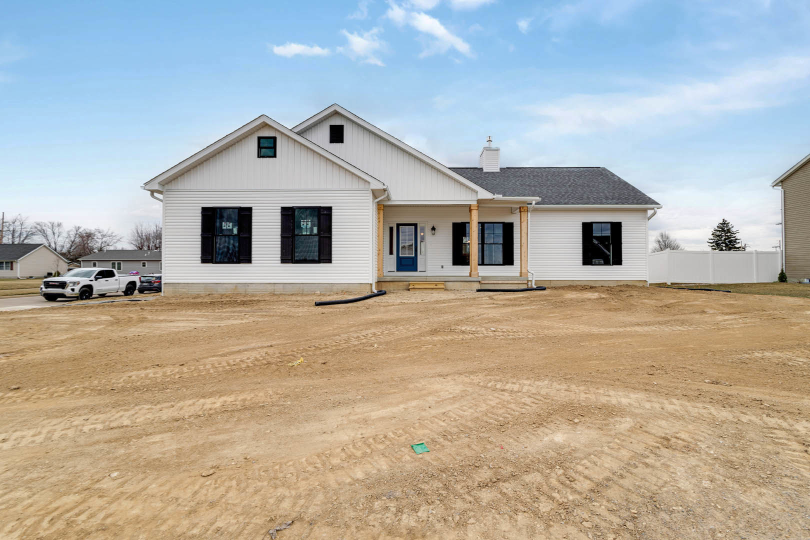 Partially built white house with blue door, open window, and leafy tree nearby; dirt lot with tire tracks and white truck under clear blue sky