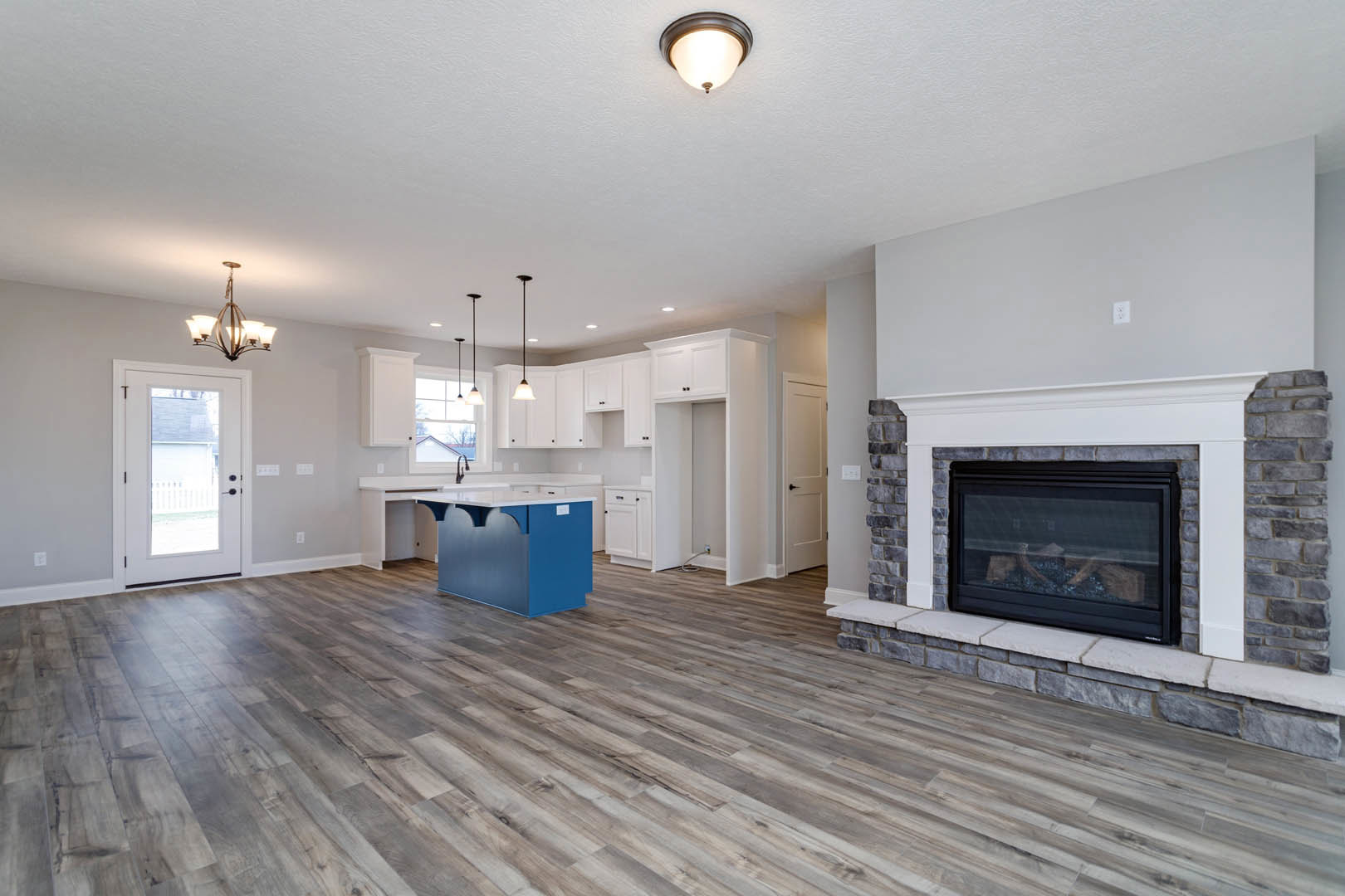 Open concept living room with white framed fireplace, wood flooring, glass windowed door, ceiling light fixture, and adjacent kitchen featuring a blue refrigerator