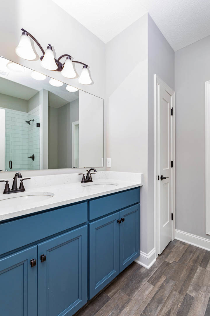 Bathroom with blue shaker cabinets, wood flooring, white countertop, large mirror, row of globe light fixtures, white door with black handle, tile shower wall, chrome faucet