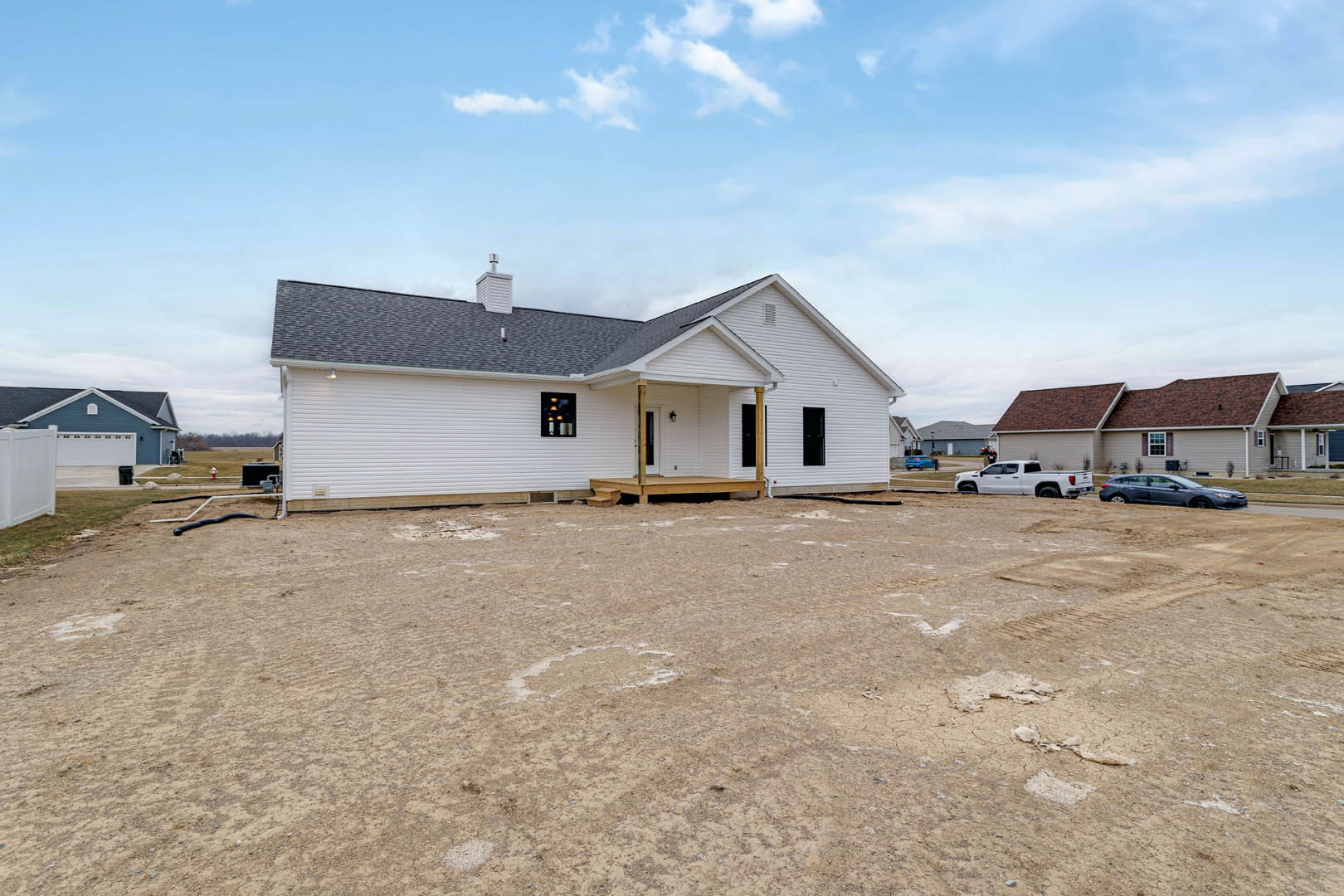 White two-story house with covered porch and tower, surrounded by dirt field, white truck parked nearby, cloudy sky overhead.