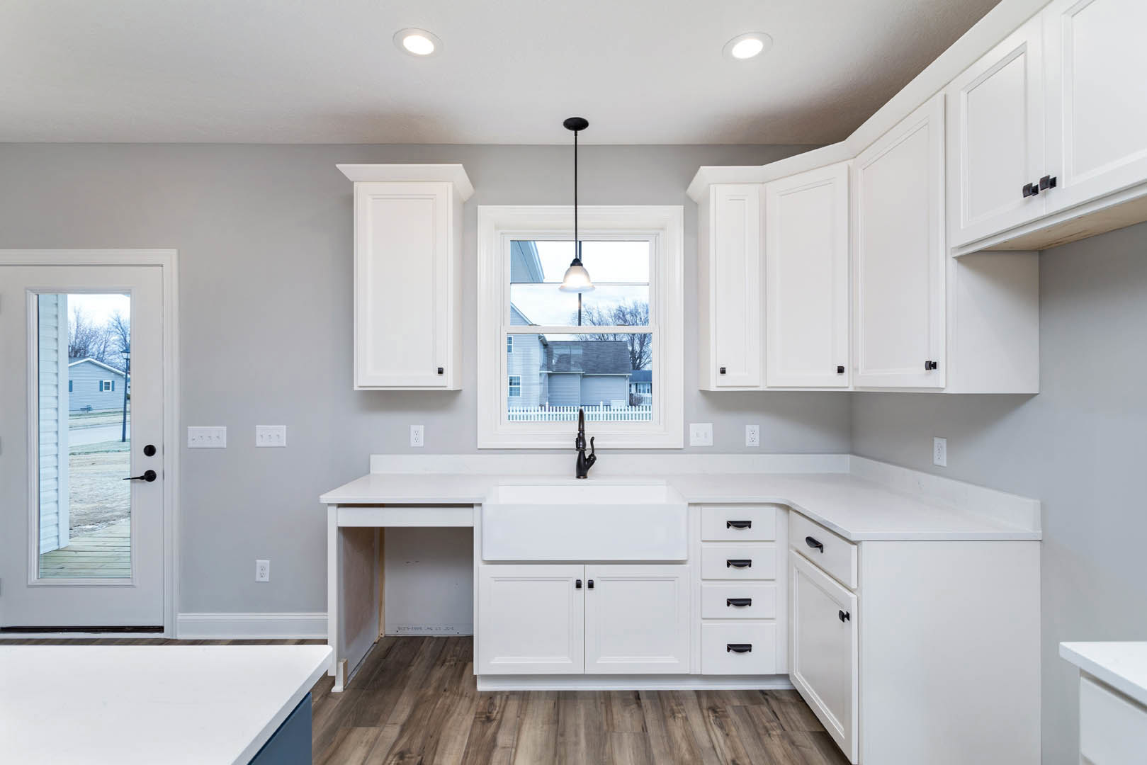 White kitchen cabinets with brushed metal handles, quartz countertops, stainless steel sink beneath a large window, and light wood flooring.