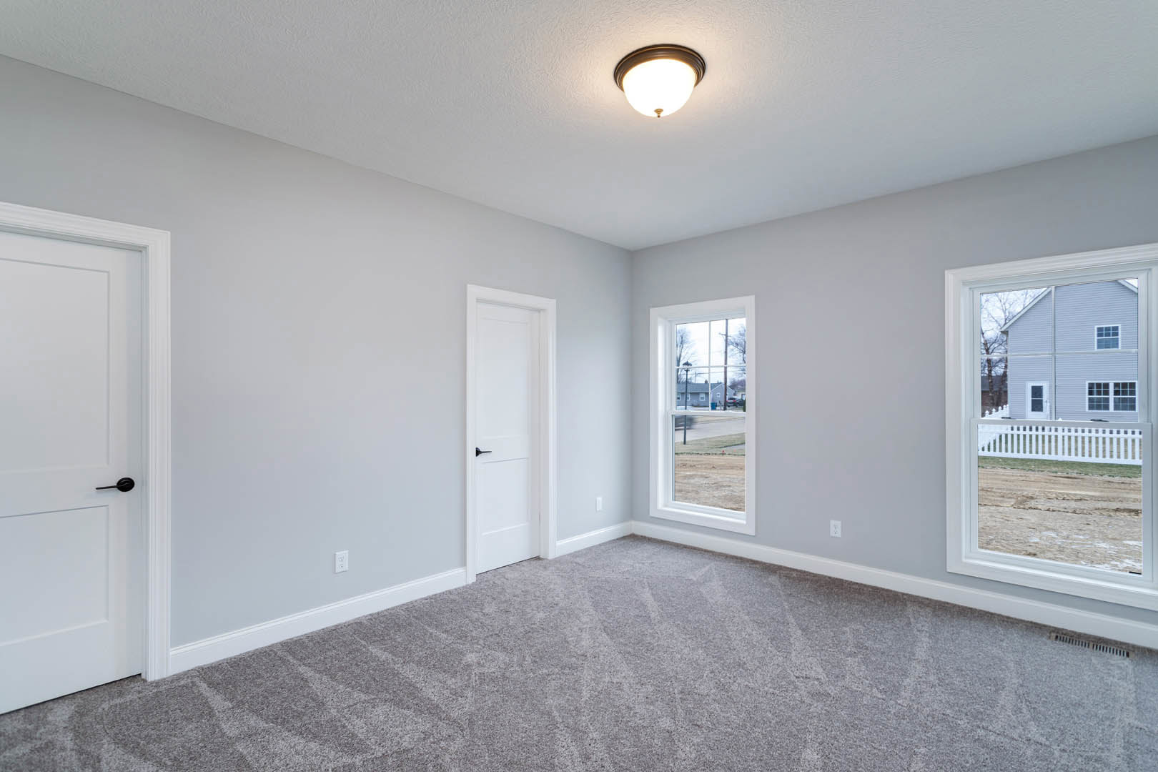 Carpeted room with white trim, white door featuring a black handle, ceiling-mounted light fixture, and windows overlooking a white fence and neighboring house
