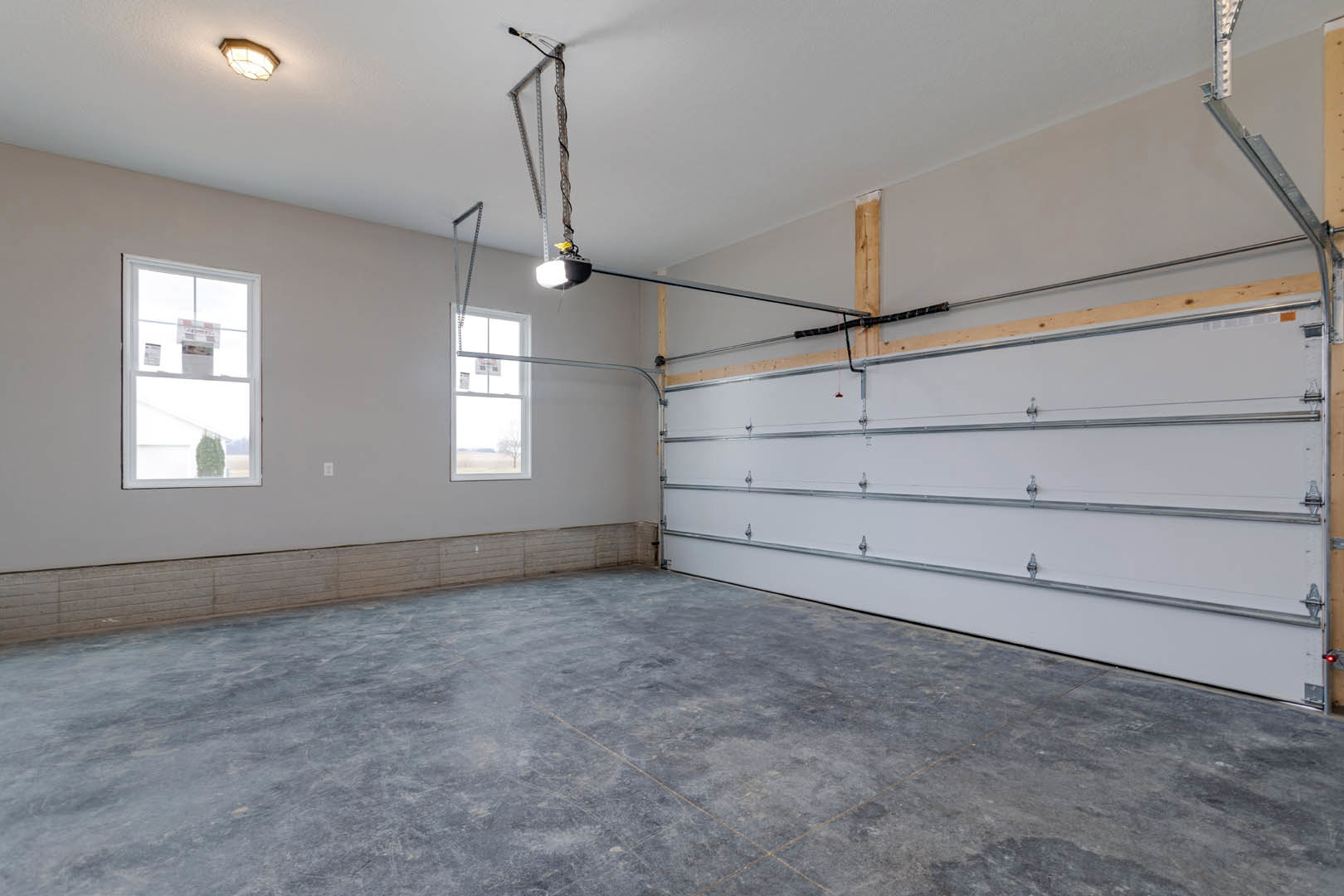 Garage interior with white plaster walls, concrete floor, ceiling-mounted light fixture, white garage door with metal bars, and window displaying a newspaper.