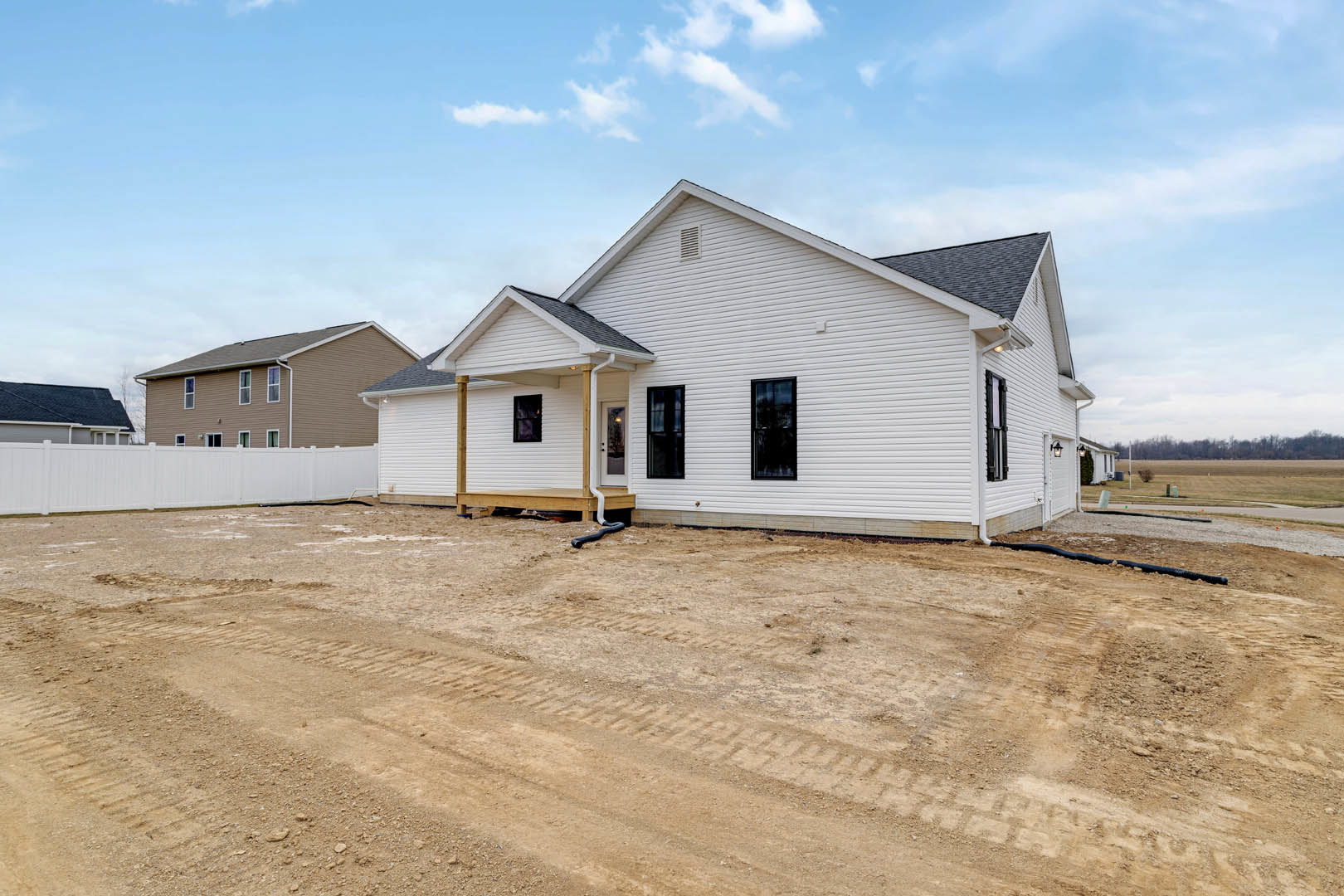 White house with black roof under construction, surrounded by dirt with tire tracks, multiple windows visible, blue sky overhead