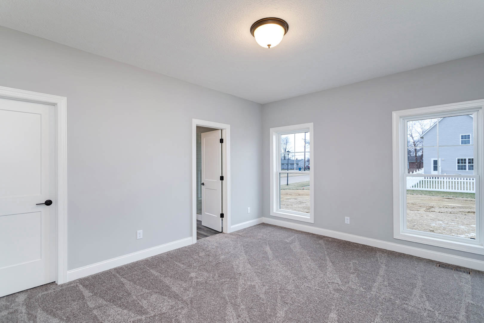 Carpeted room with white paneled door featuring black handles, ceiling light fixture, large window overlooking white fence and neighboring house, white walls with crown molding