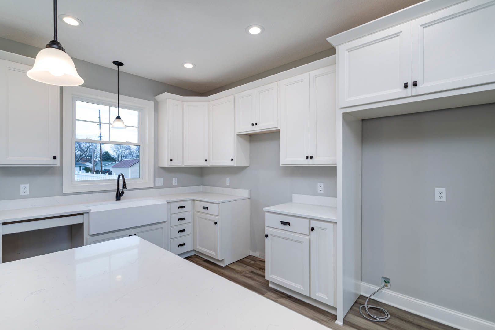 White kitchen with shaker cabinets, black hardware, quartz countertop, stainless steel faucet, window above sink, modern light fixture, and white electrical outlet with black
