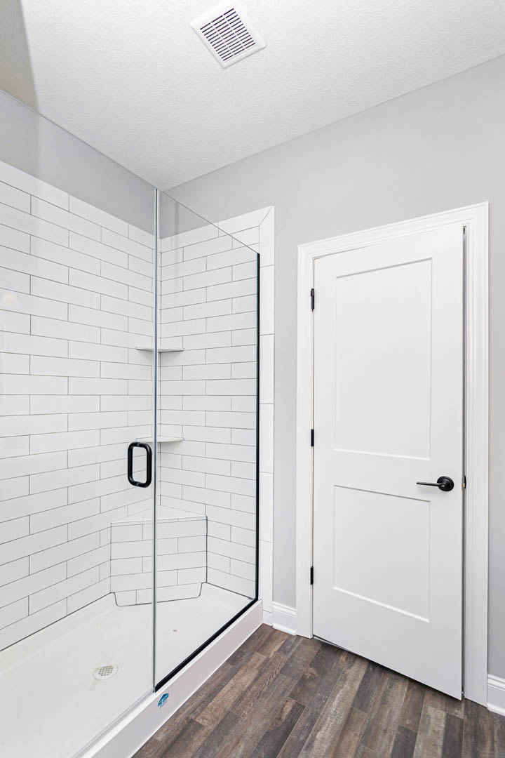 Walk-in shower with clear glass door, white walls, wood flooring, white door with black handle, and rectangular white vent with black lines