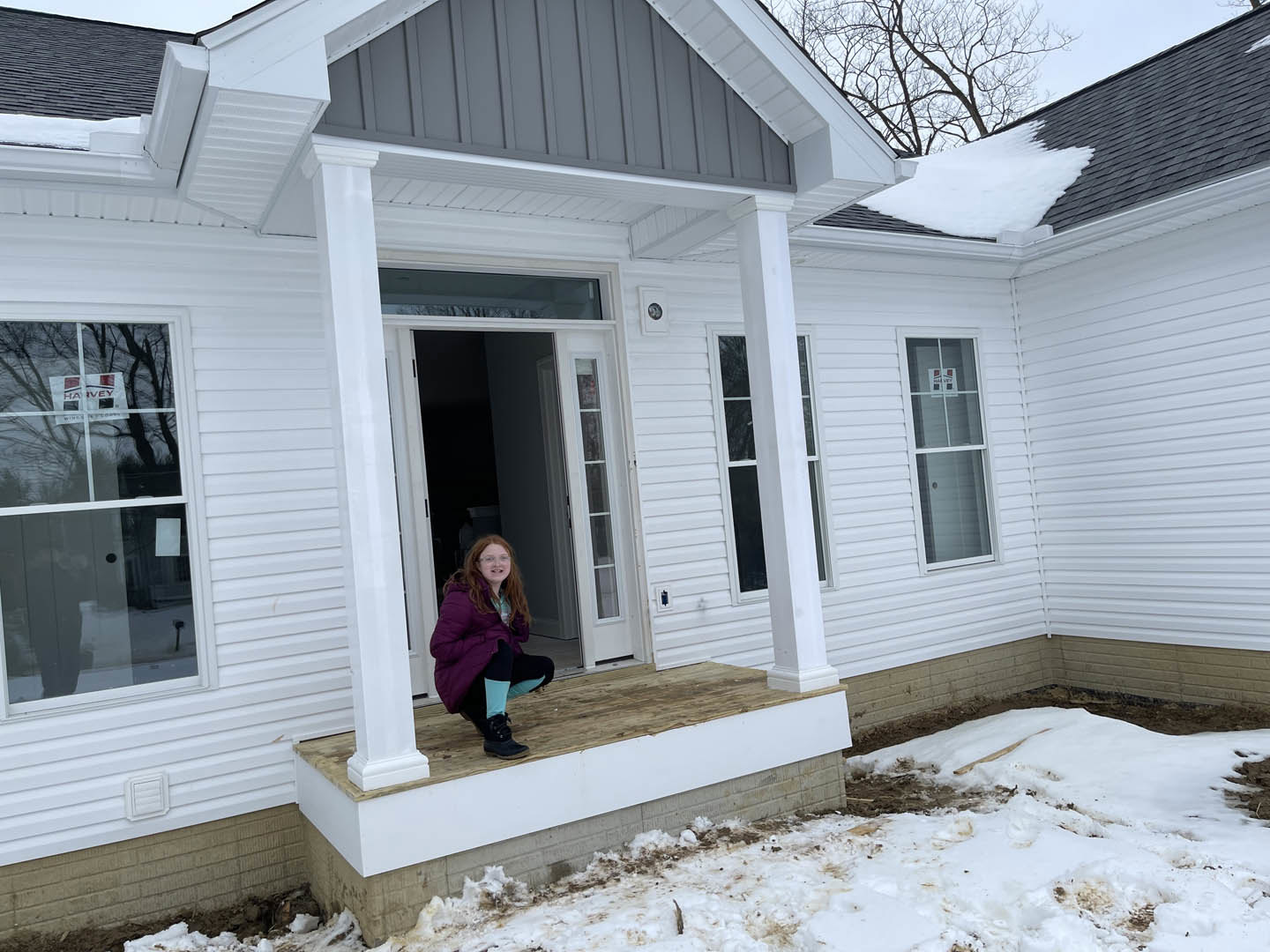 Girl in a purple coat sitting on a porch with white siding, red-lettered sign, windows including one with a cross, winter outdoor setting