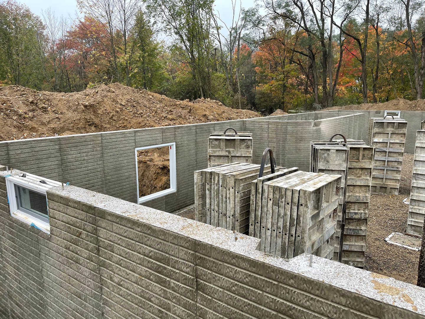 Construction site with stacked cement blocks, brick wall with window, metal container, dirt ground, and trees in the background