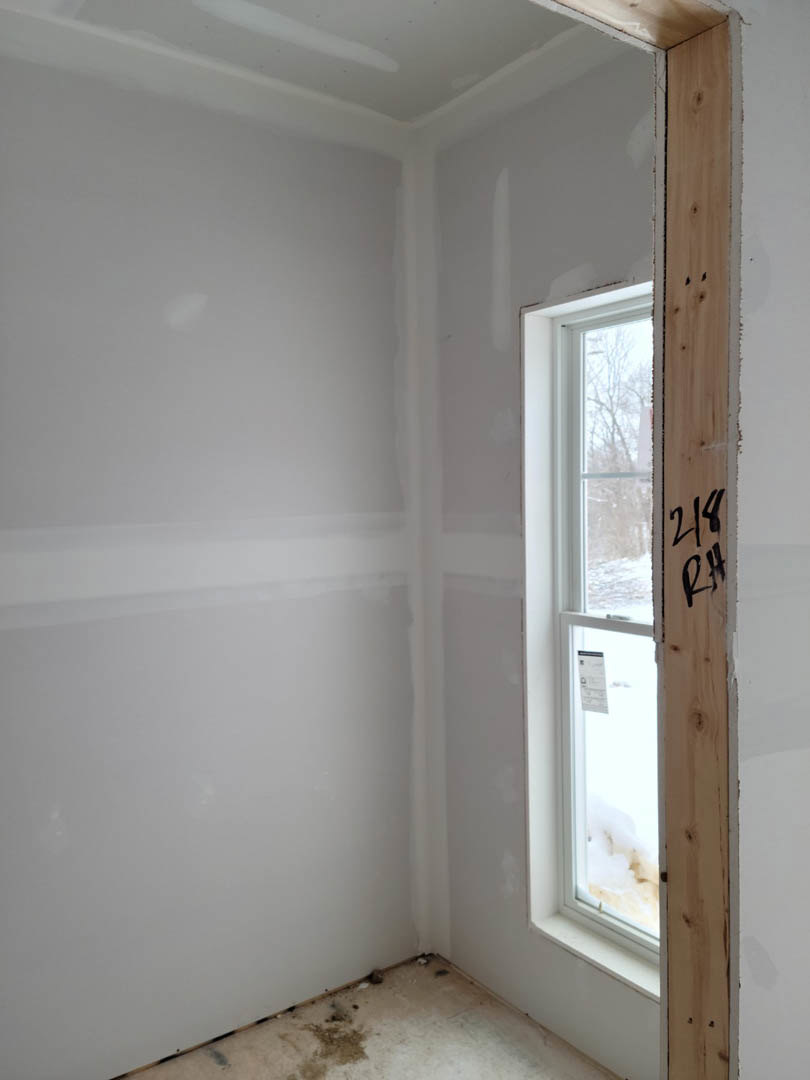 Room with plaster walls, wood-framed window, and unfinished floor with visible dirt; close-up of numbers marked on wood surface and a white object near window displaying a sign.