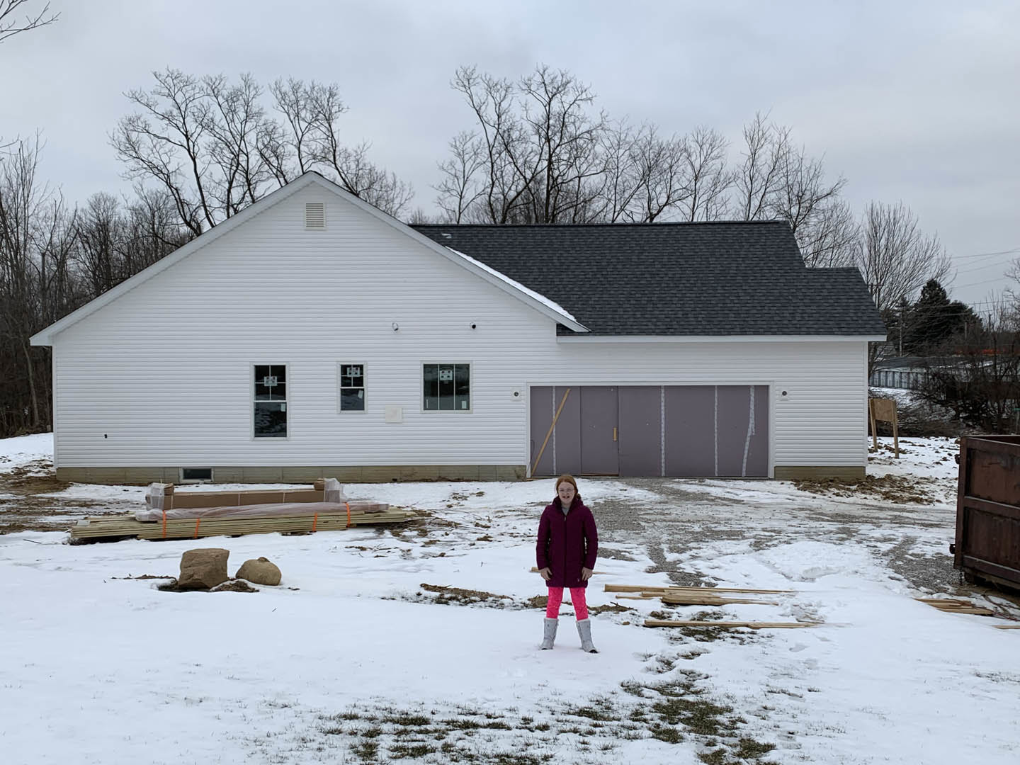 White house with attached garage, grey front door with white trim, snow-covered yard, large rock in foreground, girl in pink pants and coat standing outside, bare trees and cloudy