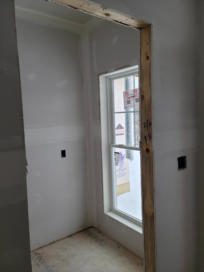 Sunlit room featuring a large window with wood trim, smooth plaster walls, light-colored flooring, and a visible wall opening.