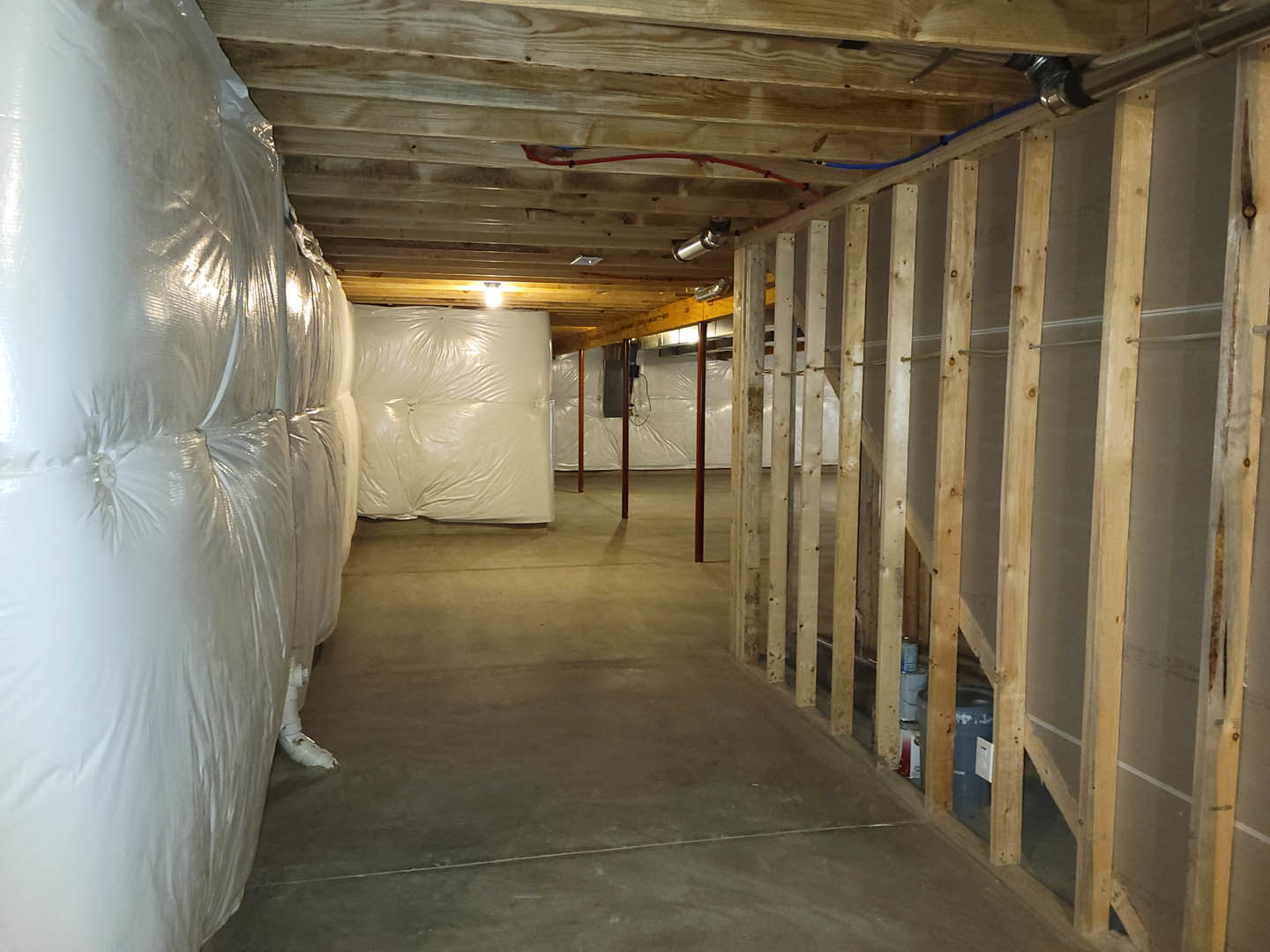 Bedroom with exposed wood beams, wood-paneled wall, white plastic bags on bed, white pillow, visible pipe, and unfinished surfaces.