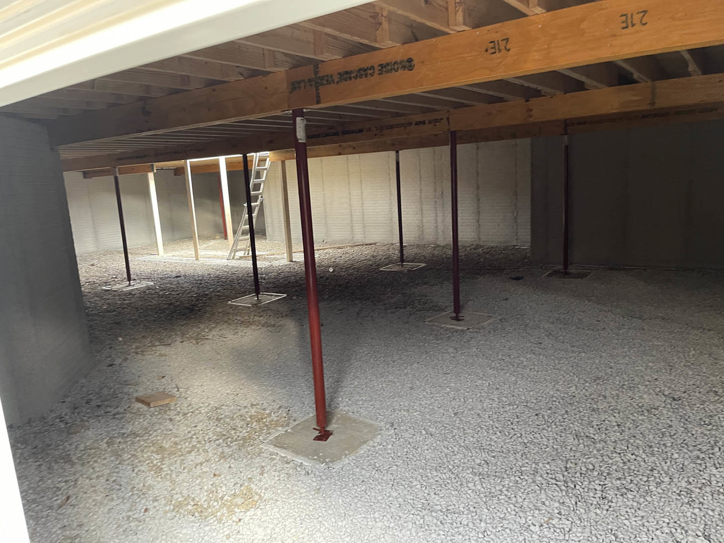 Concrete floor with a red metal pole, wooden ceiling beam marked with black writing, and a ladder leaning against an unfinished wall in an indoor room.