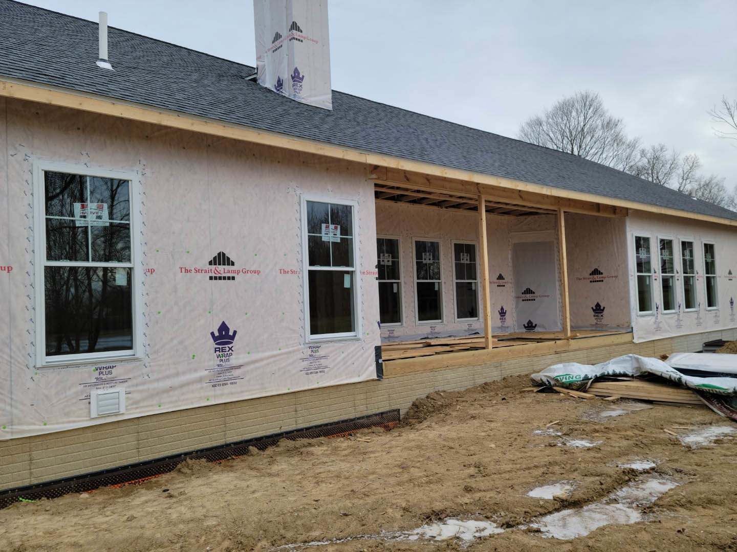 Two-story house under construction with exposed framing, covered front porch, white vent on exterior wall, dirt yard, and window displaying a construction sign.