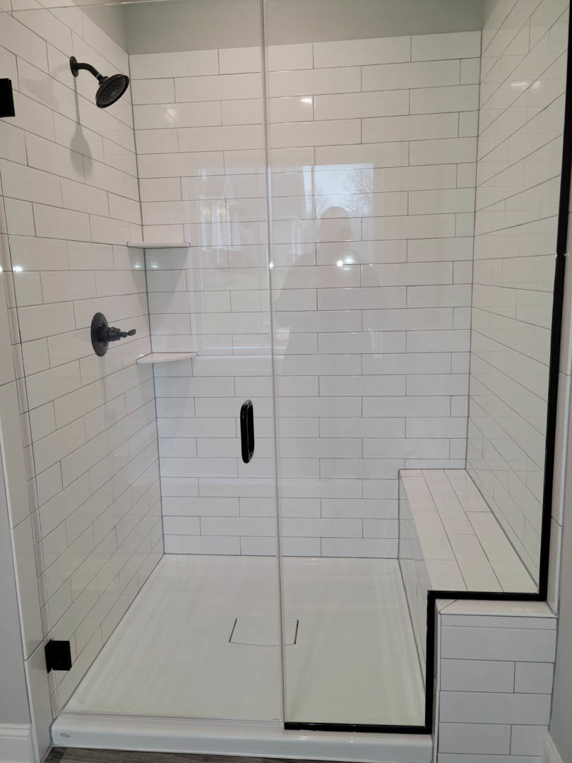 Walk-in shower with clear glass door, matte black shower head and pole, white tiled walls and floor, square drain, white vanity with matching countertop