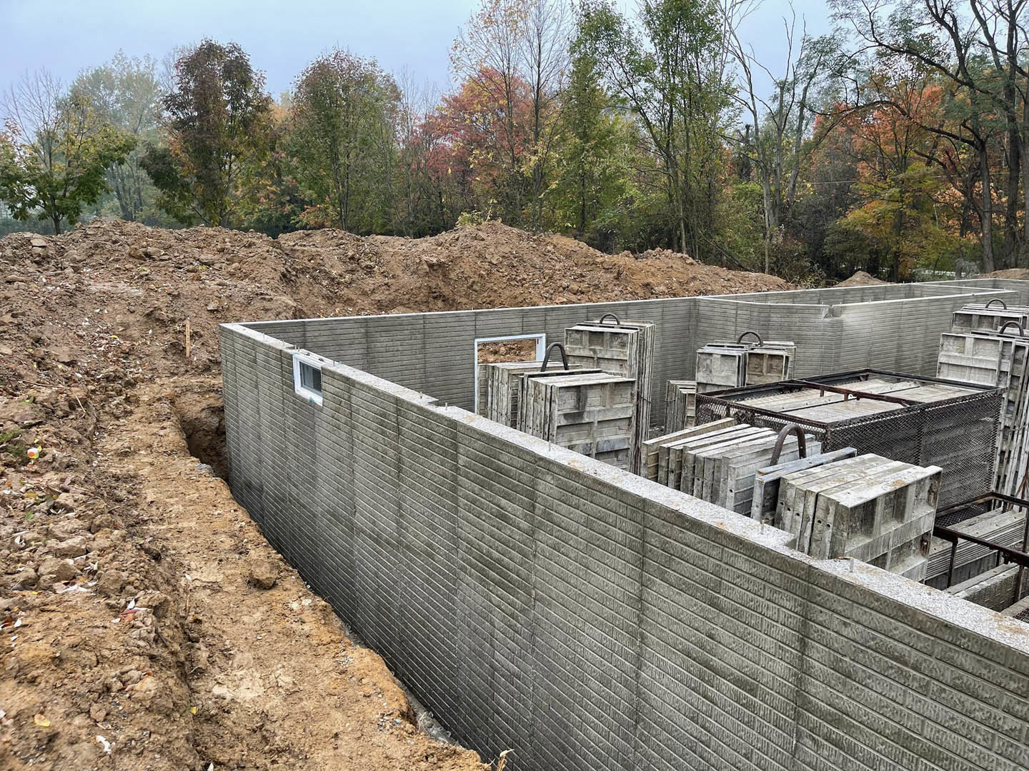Concrete foundation wall with exposed hole in the ground, surrounded by dirt, stacked concrete blocks, and trees with orange and green leaves