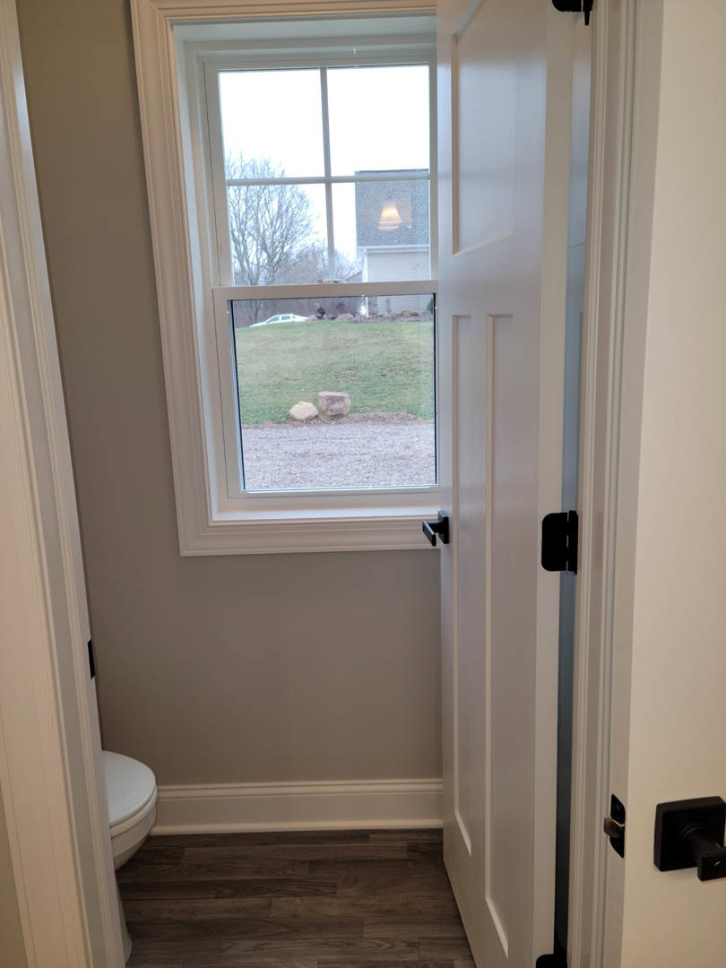 Bathroom with wood flooring, white baseboards, large window overlooking grassy yard, black door handle, neutral walls, and modern fixtures.