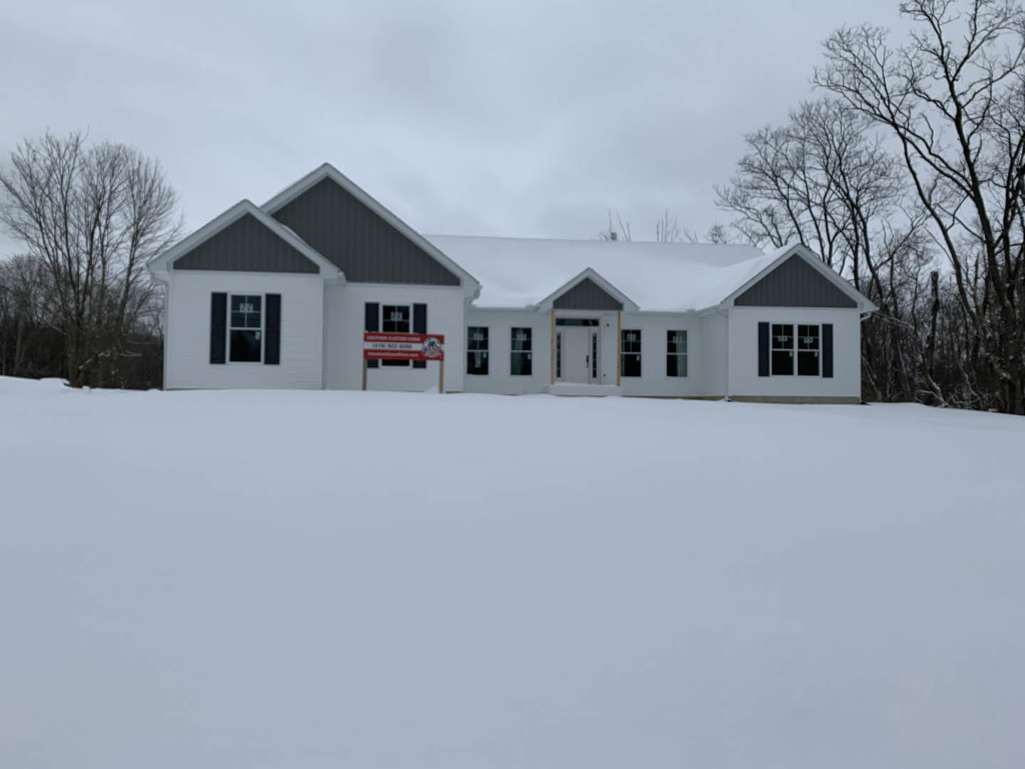 Two-story home with white siding and dark roof, snow covering ground and roof, leafless trees in background, windows reflecting overcast sky, blurry red sign near entrance
