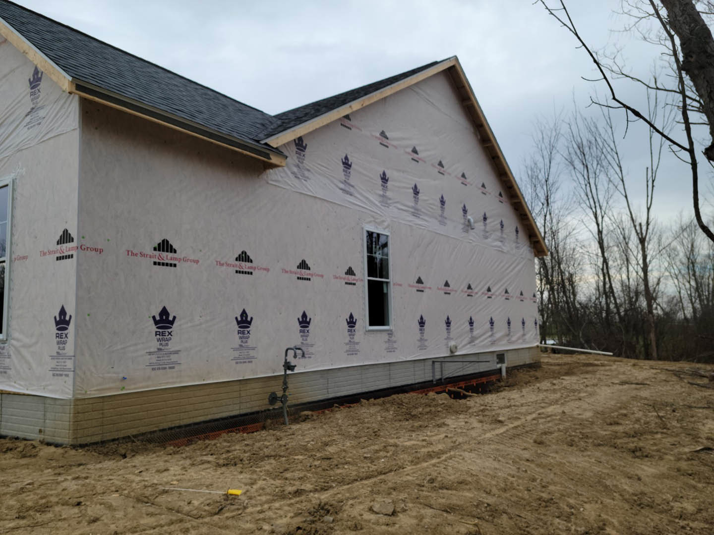 White plastic sheeting draped over the side of a house under construction, white-framed window visible, tree branches in foreground, exposed dirt with water faucet, cloudy sky