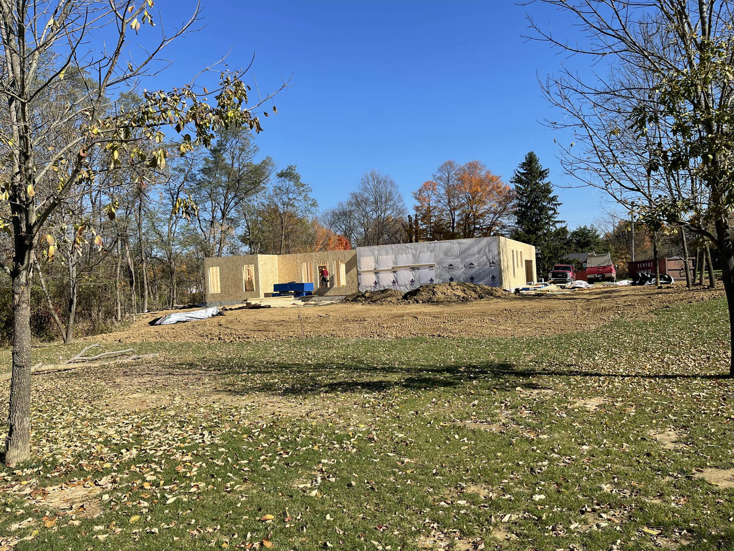 Framed custom home under construction on grassy lot, surrounded by trees, under clear blue sky