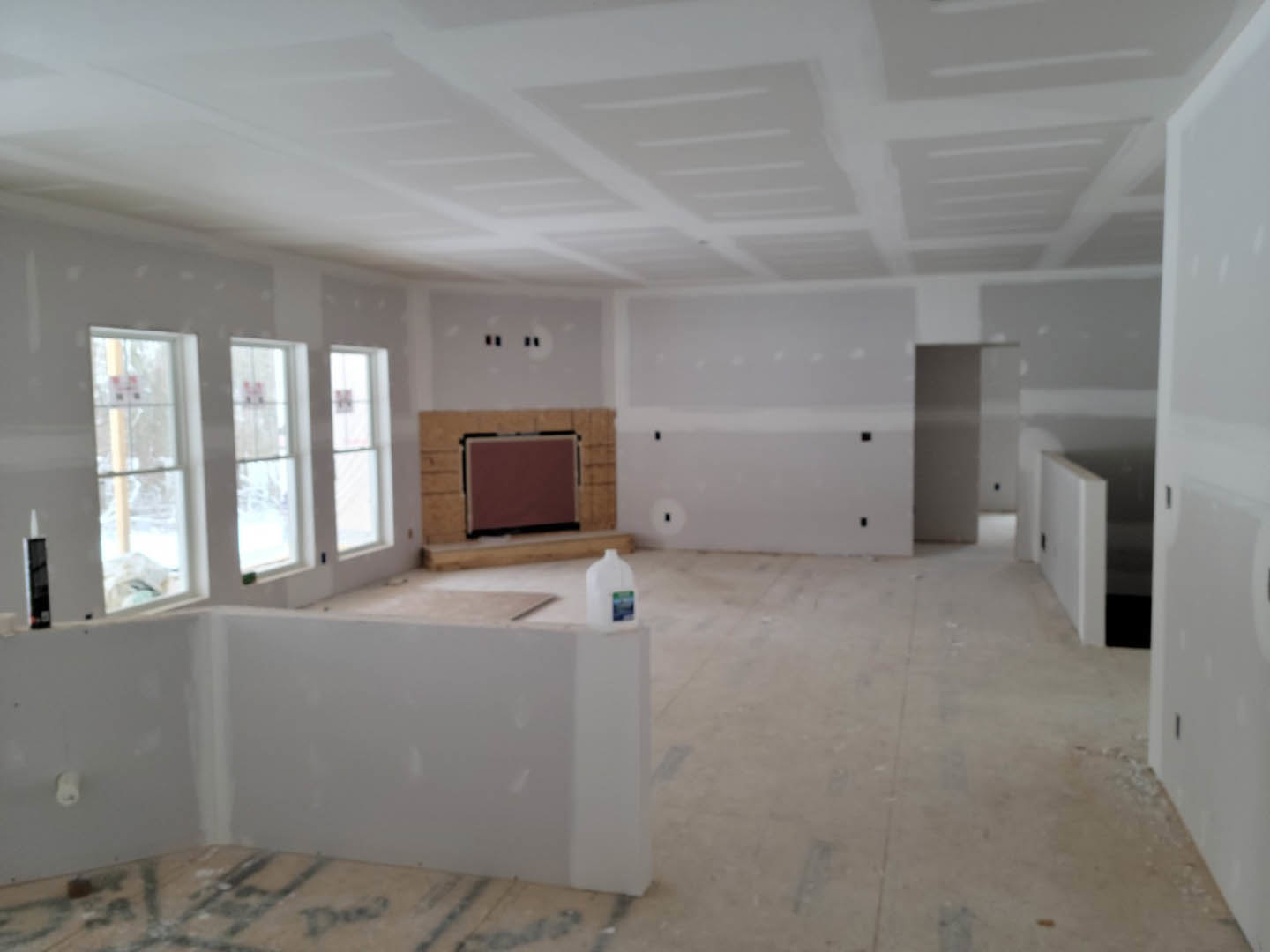 Living room with stone fireplace, white plaster walls, hardwood flooring, and a row of large windows letting in natural light