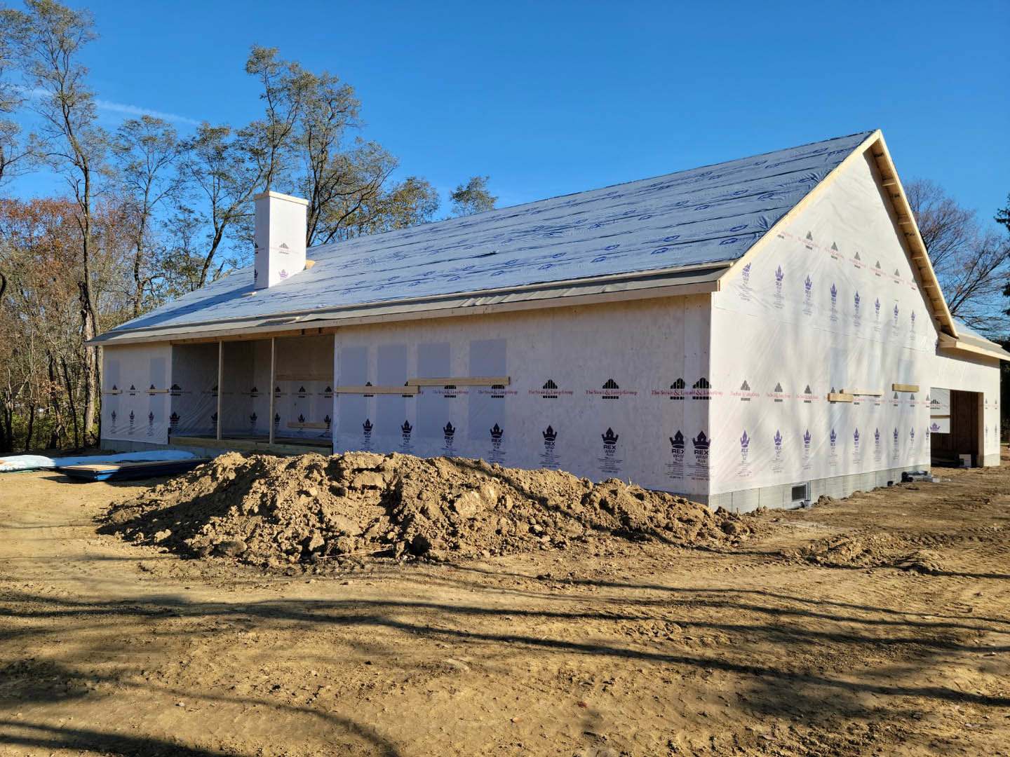 Framed house under construction with exposed wooden beams, piles of dirt in foreground, surrounding trees, and clear blue sky