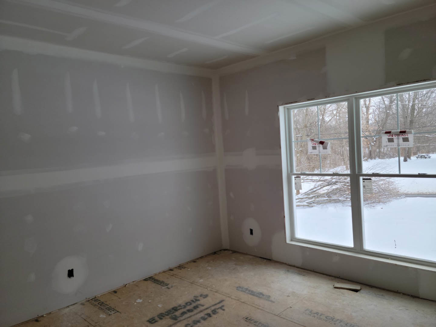Unfinished room with two windows, snow visible outside, plaster walls with holes, exposed ceiling, and close-up of light-colored floor; red and white striped sign and labeled box
