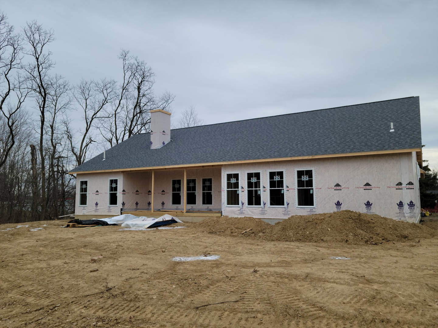 Two-story house under construction with multiple white-framed windows, white pillars marked with red tape, surrounded by dirt piles and mature trees