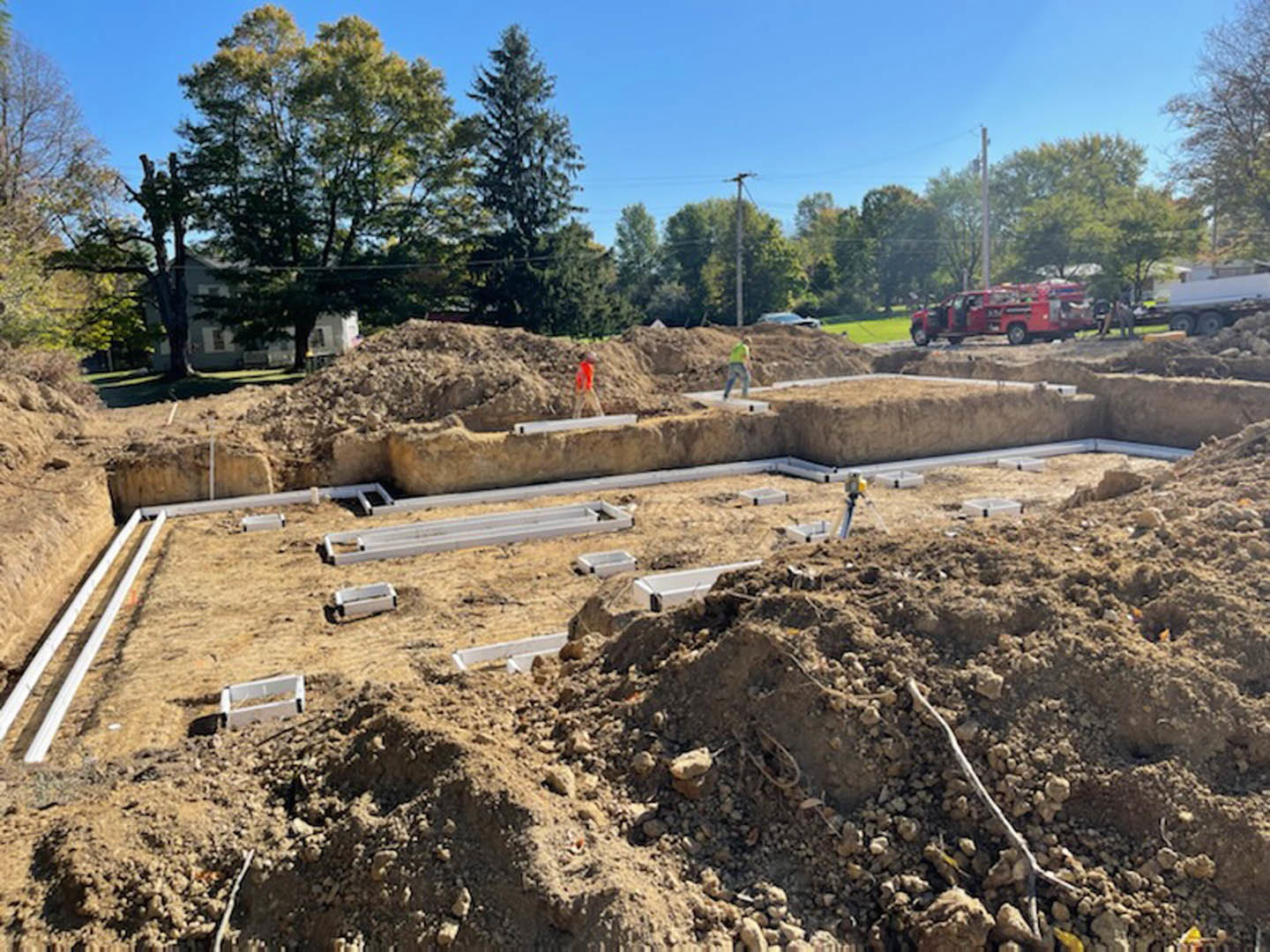 Construction site with workers, red truck with open doors, pile of dirt and metal beams, tree and telephone poles, trailer carrying another trailer, cloudy sky overhead
